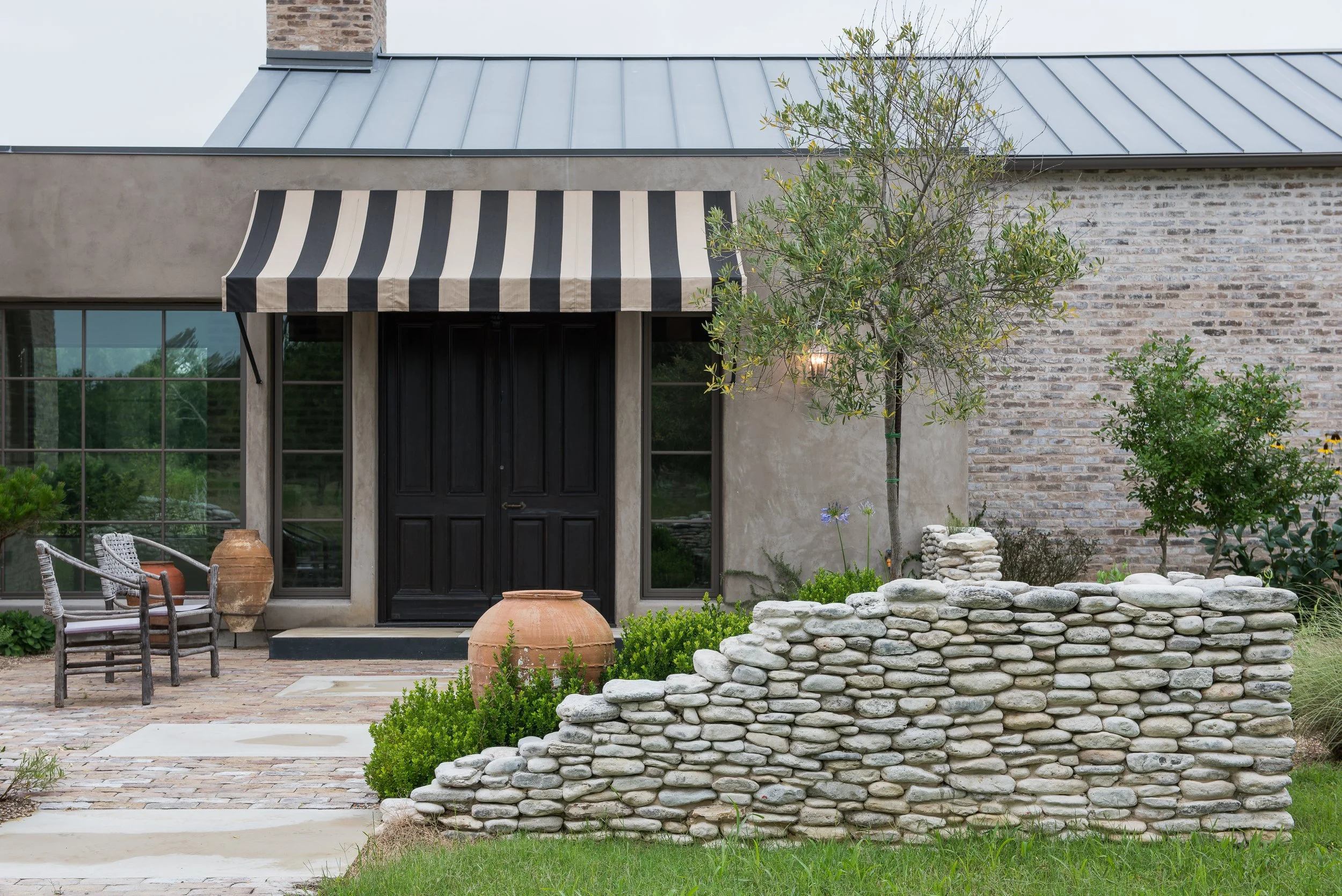 Exterior of unique ranch house with a black door, striped awning, patio furniture, large terracotta pots, a stone wall with plants, and a landscaped yard with trees and shrubs.