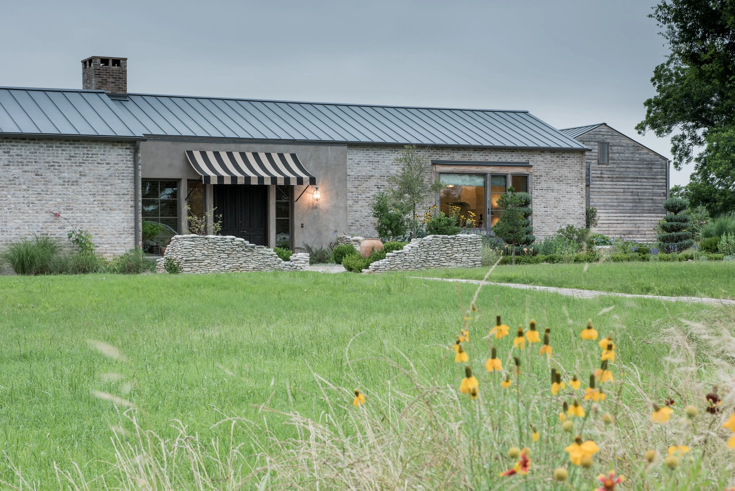Eclectic house with a gray metal roof and brick exterior, surrounded by a lush green lawn and garden with various plants and yellow flowers, under a clear sky.
