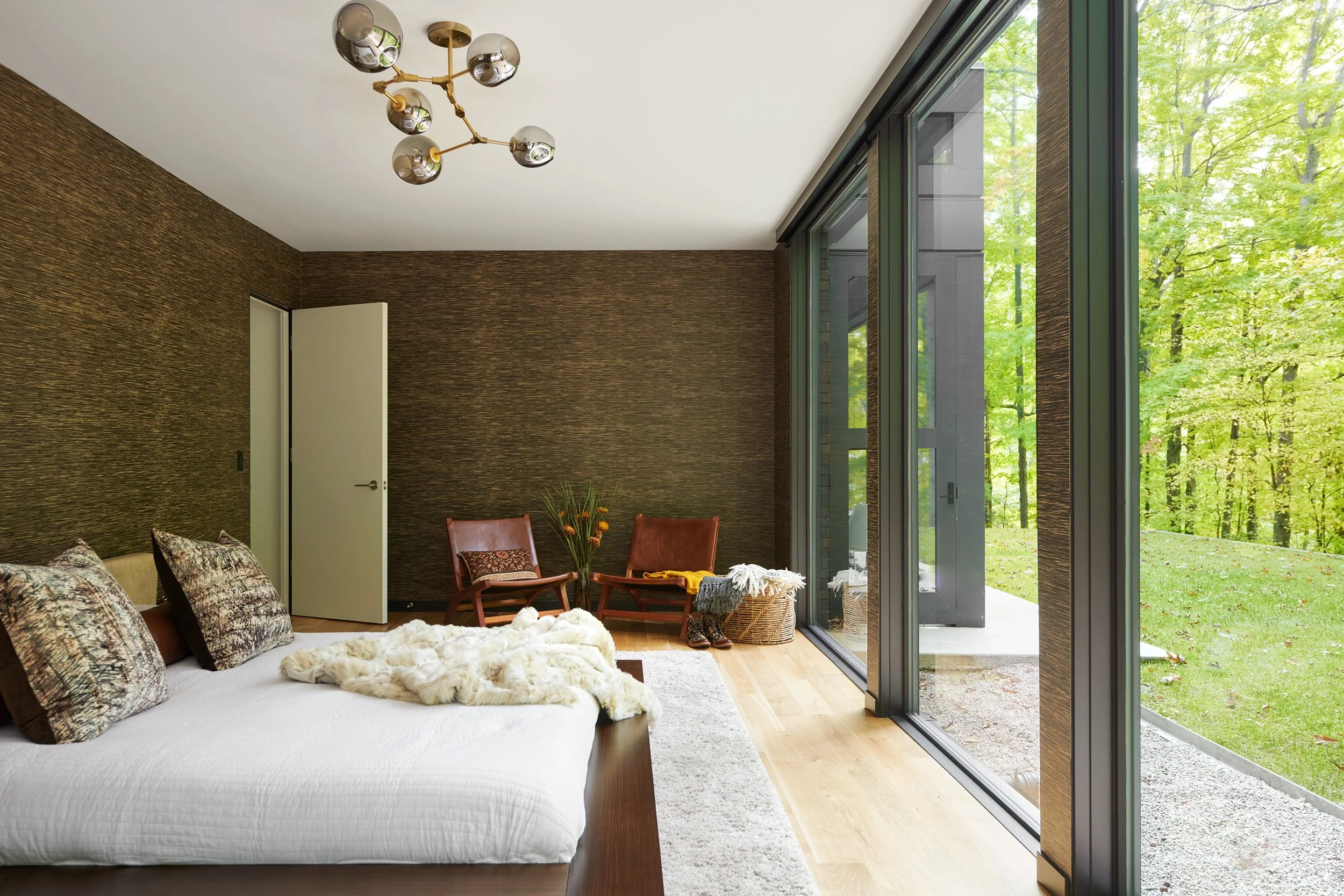 Modern bedroom with large glass windows overlooking green trees, a white bed with patterned pillows and a fur blanket, two red armchairs, a small table with a vase of flowers, and a dark textured accent wall.