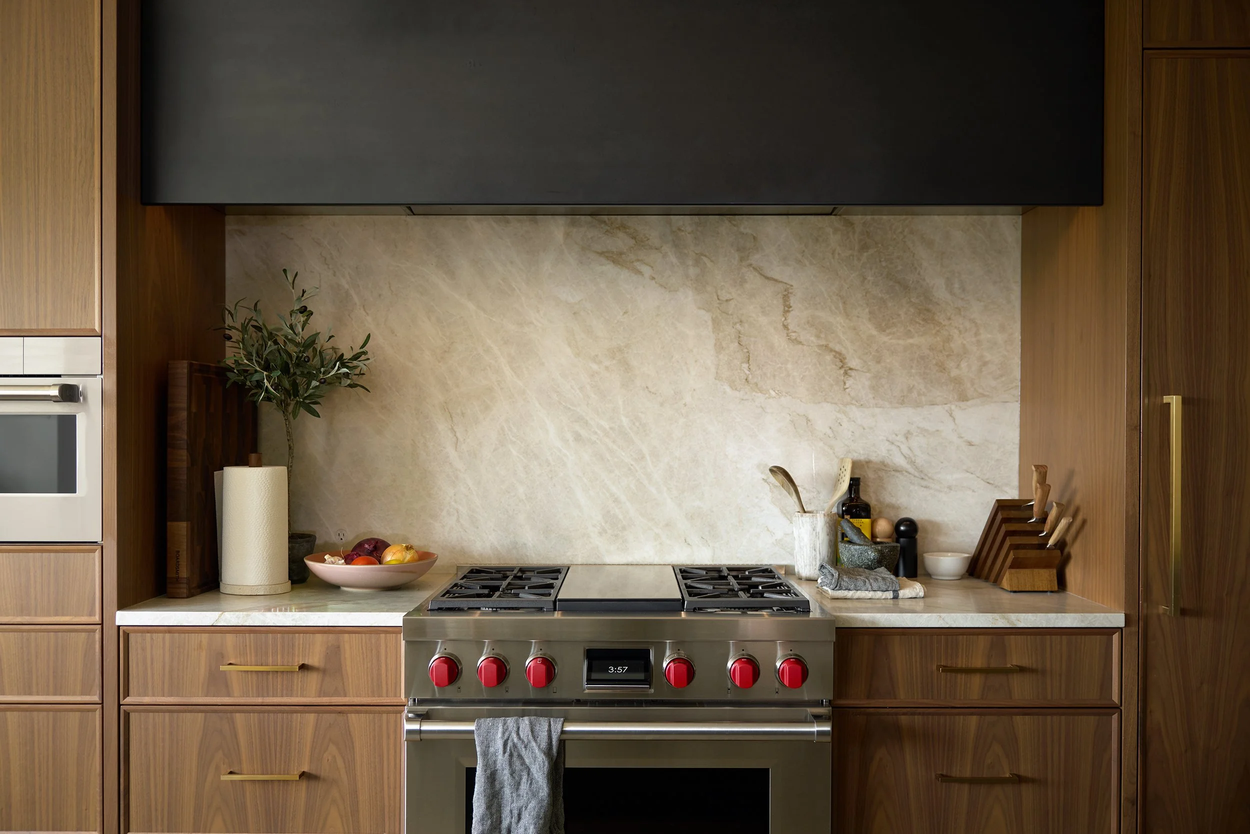 Kitchen inset: oven, range with bright red knobs, and beige quartz backsplash with custom black hood