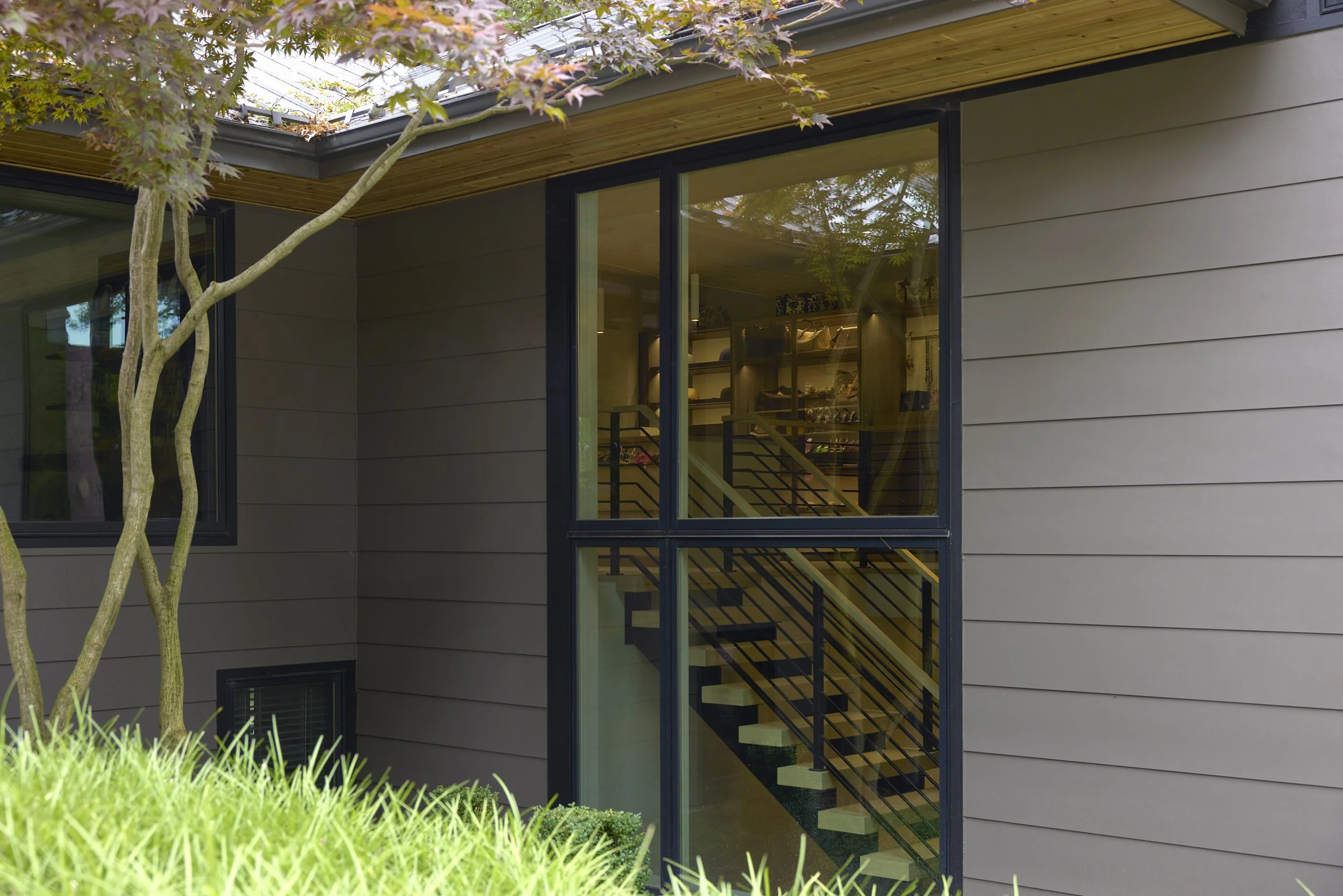Exterior view of a modern lakefront home addition with gray siding and large black-framed glass windows, showing a staircase inside through the window, with trees and green grass in the foreground.