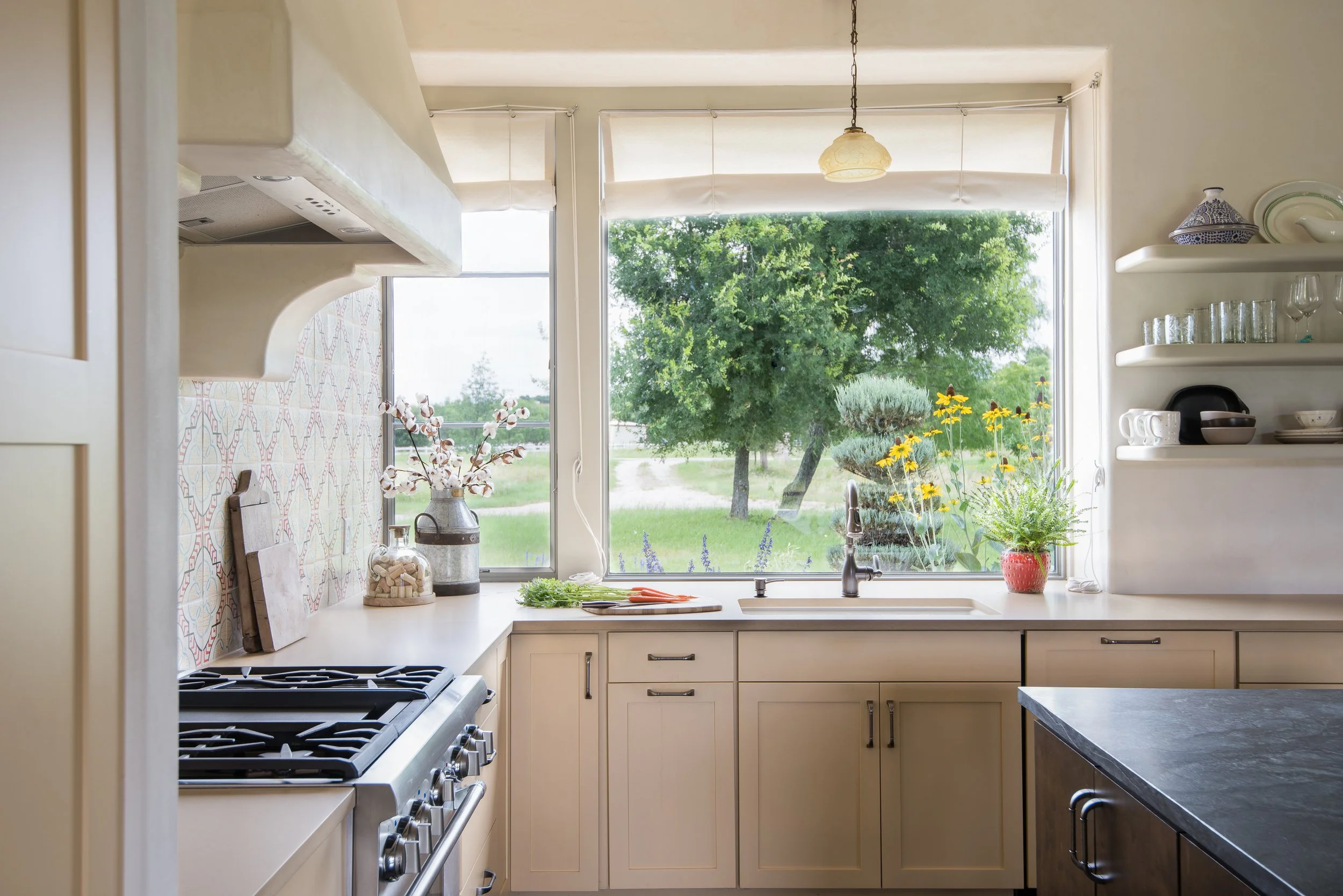A bright kitchen with cream cabinets, a window with a view of green trees, a stove on the left, and open shelves with glassware on the right.