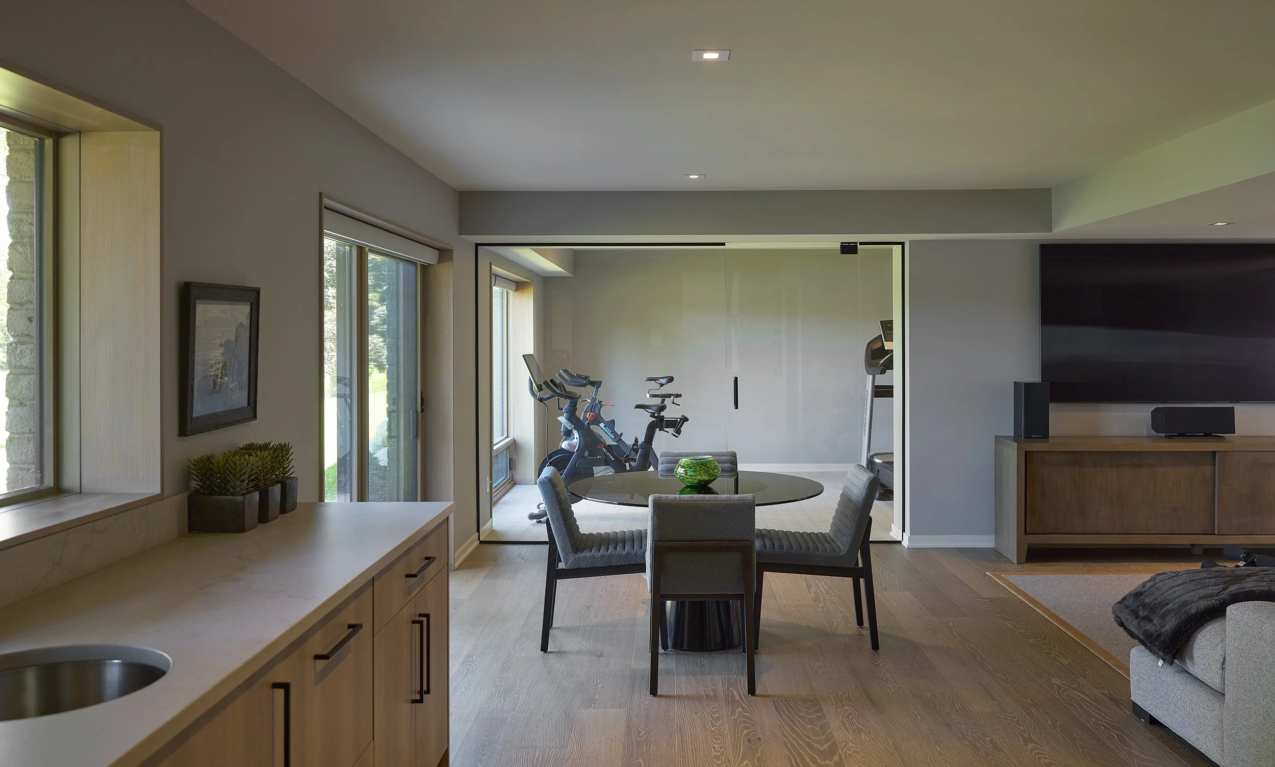 Lounge with bar and dining table, home gym in background with stationary exercise bike, treadmill, and large flat-screen TV on a wooden console.