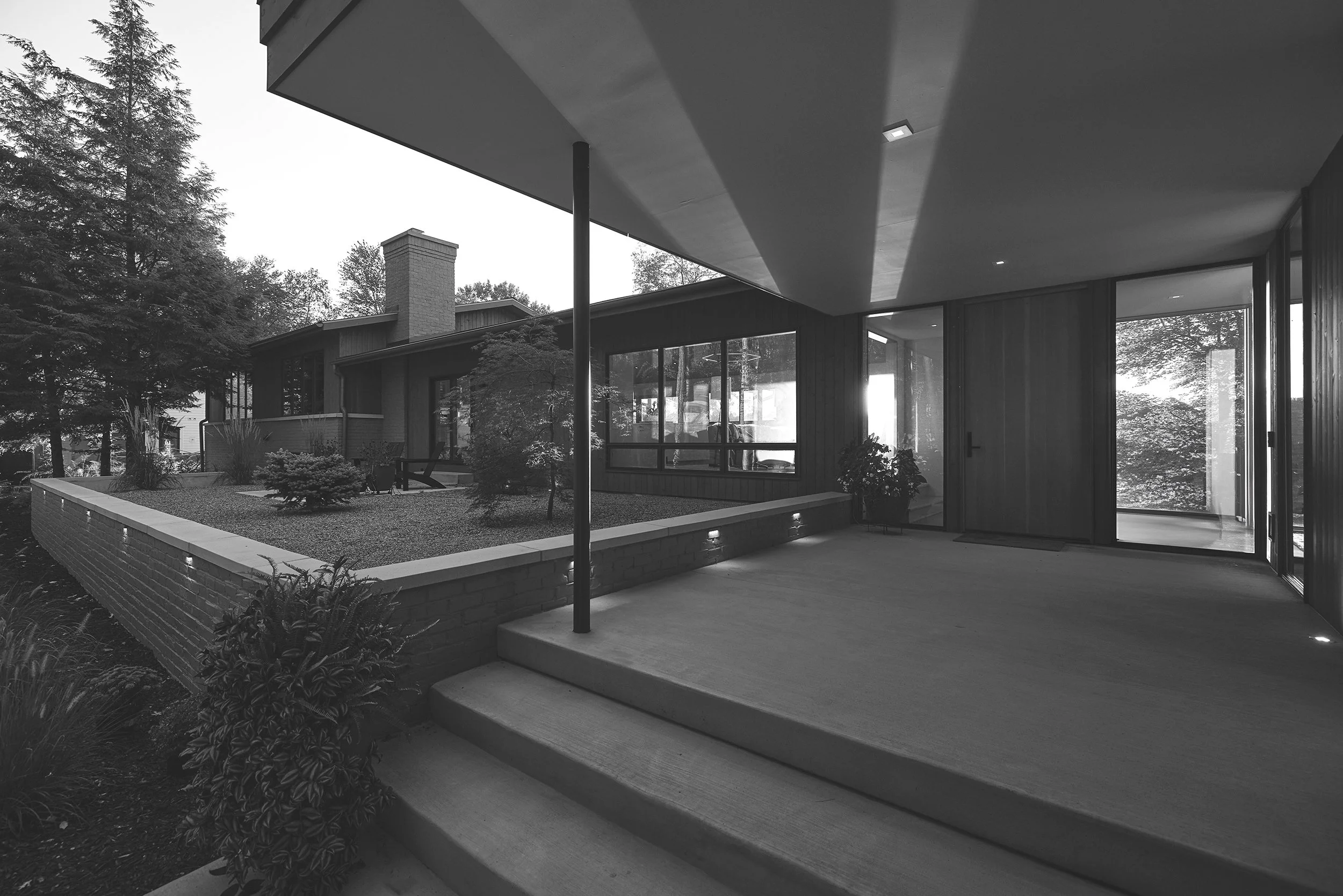 Black and white photo of front entryway of modern house with steps leading to a custom wood door, large windows, landscaped yard and surrounding trees