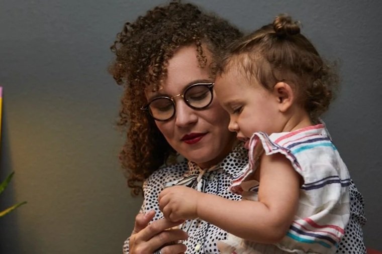 A woman with curly hair and glasses holding a young girl with a top knot in her hair, both smiling and looking at an object in the woman's hand.