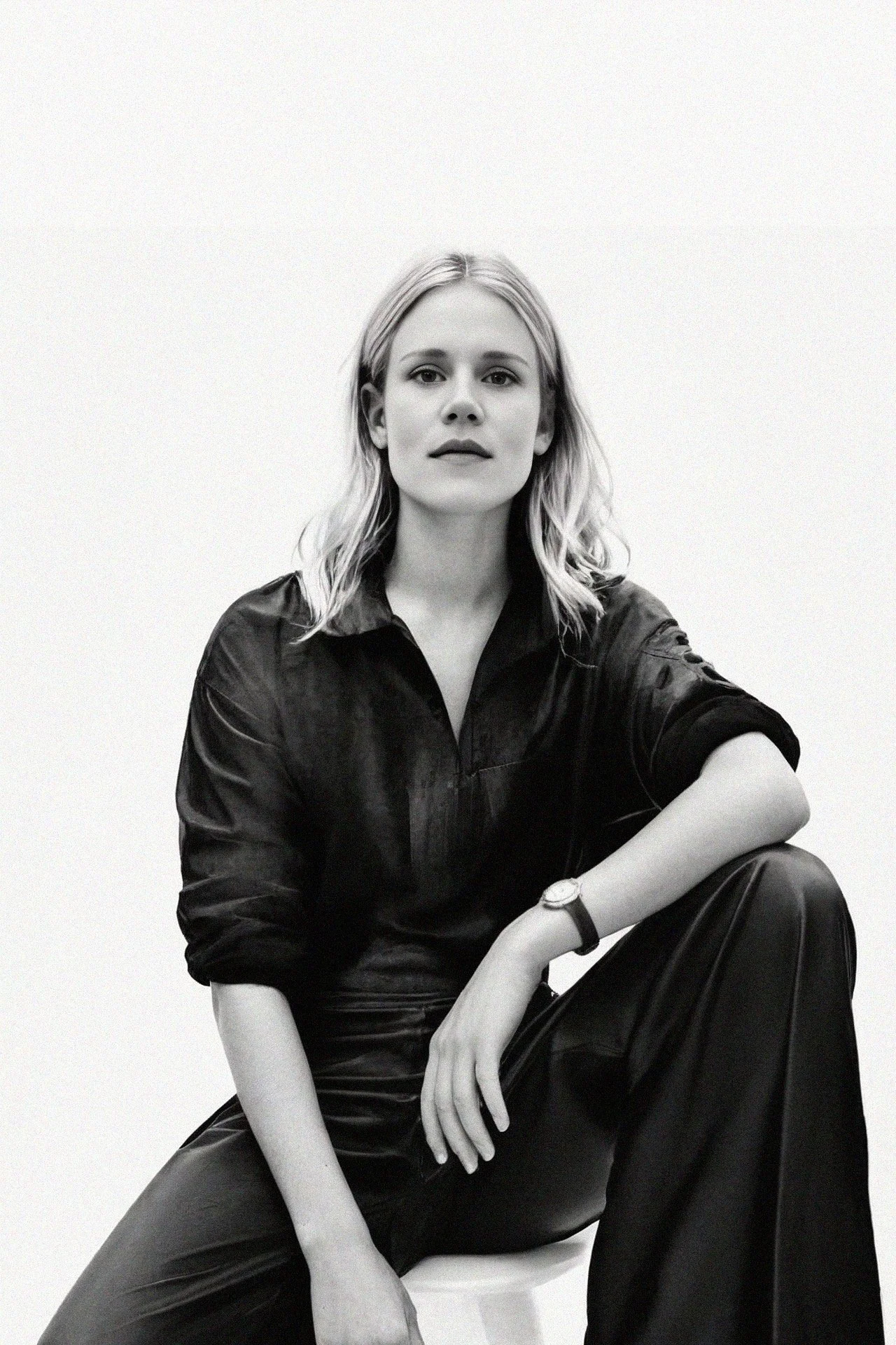 A woman with shoulder-length blonde hair, wearing a black leather shirt and black leather pants, sitting against a plain white background with her legs crossed and her left arm resting on her knee.