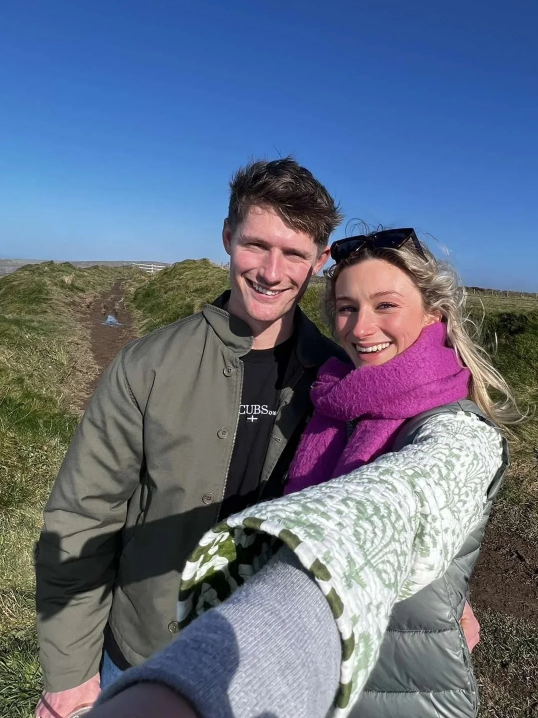 A smiling young man and woman taking a selfie outdoors on a sunny day, standing on a grassy and dirt trail with a clear blue sky in the background.