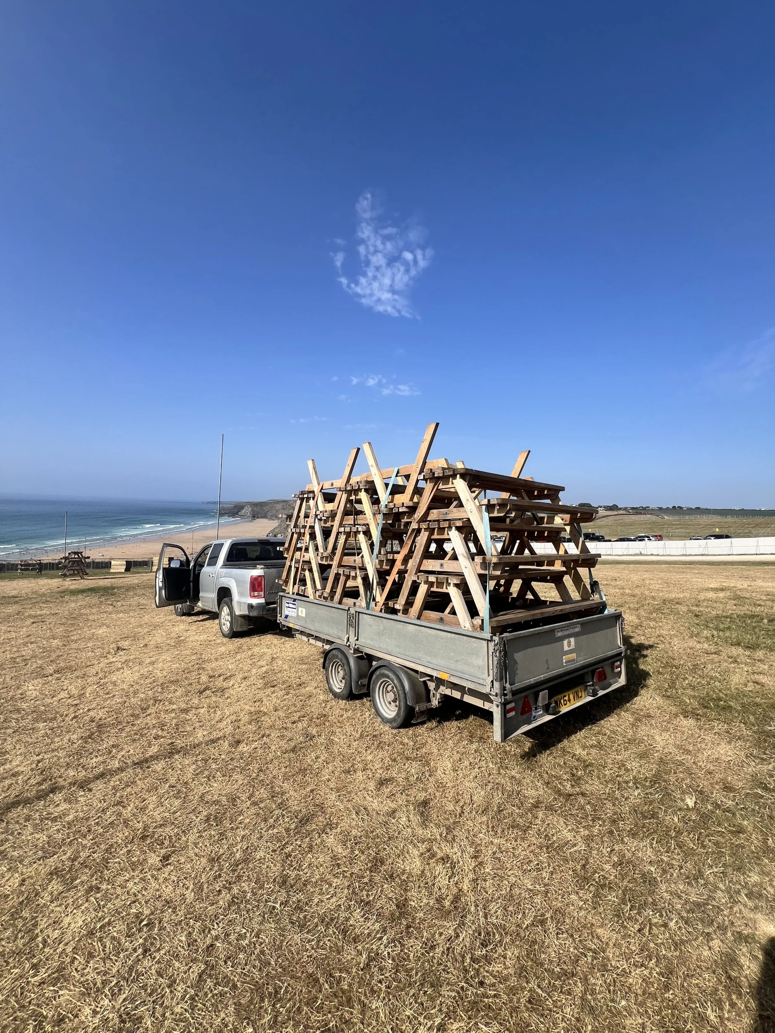 A pickup truck towing a trailer filled with wooden picinic benches, parked on a grassy field near a beach with cliffs and a blue sky.