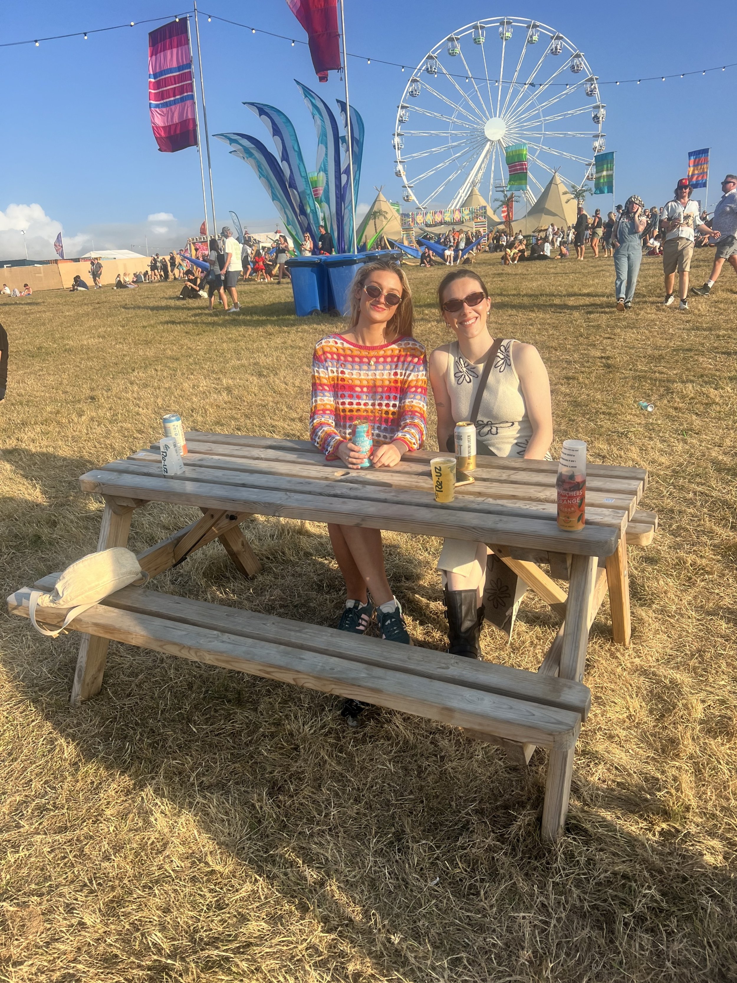Two women sitting at a wooden picnic table with cans of drinks, smiling at the camera, at a fairground with a Ferris wheel and tents in the background, under a clear blue sky.