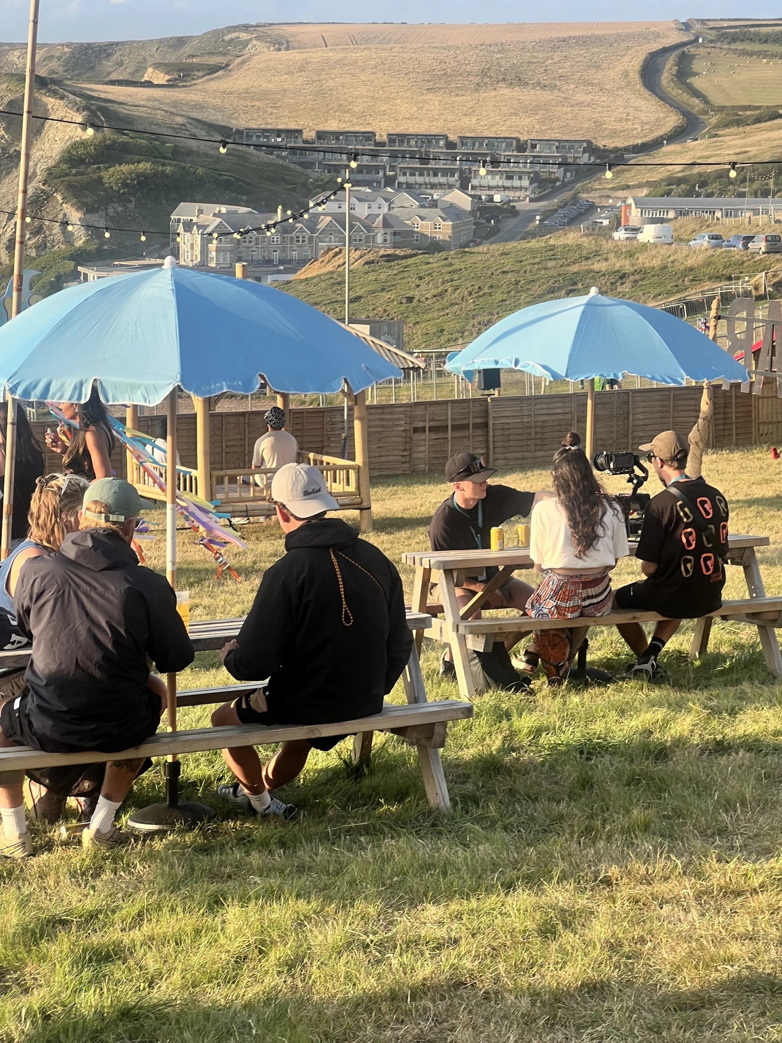 People sitting at picnic tables under blue umbrellas in a grassy outdoor area with a camera crew filming a woman, with houses and hills in the background.