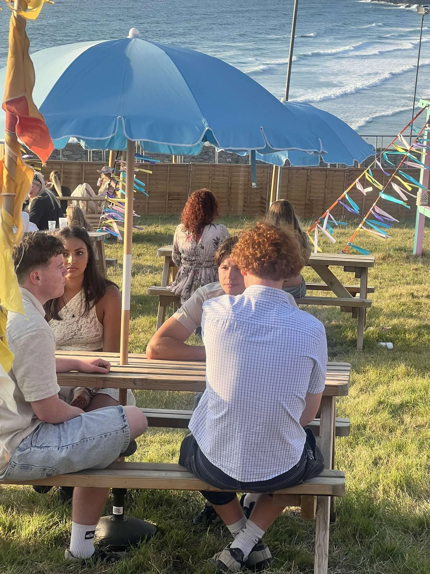 Young people sitting at a picnic table outdoors near the beach, some under a blue umbrella, with colorful decorations in the background. At a Cornish Festival.