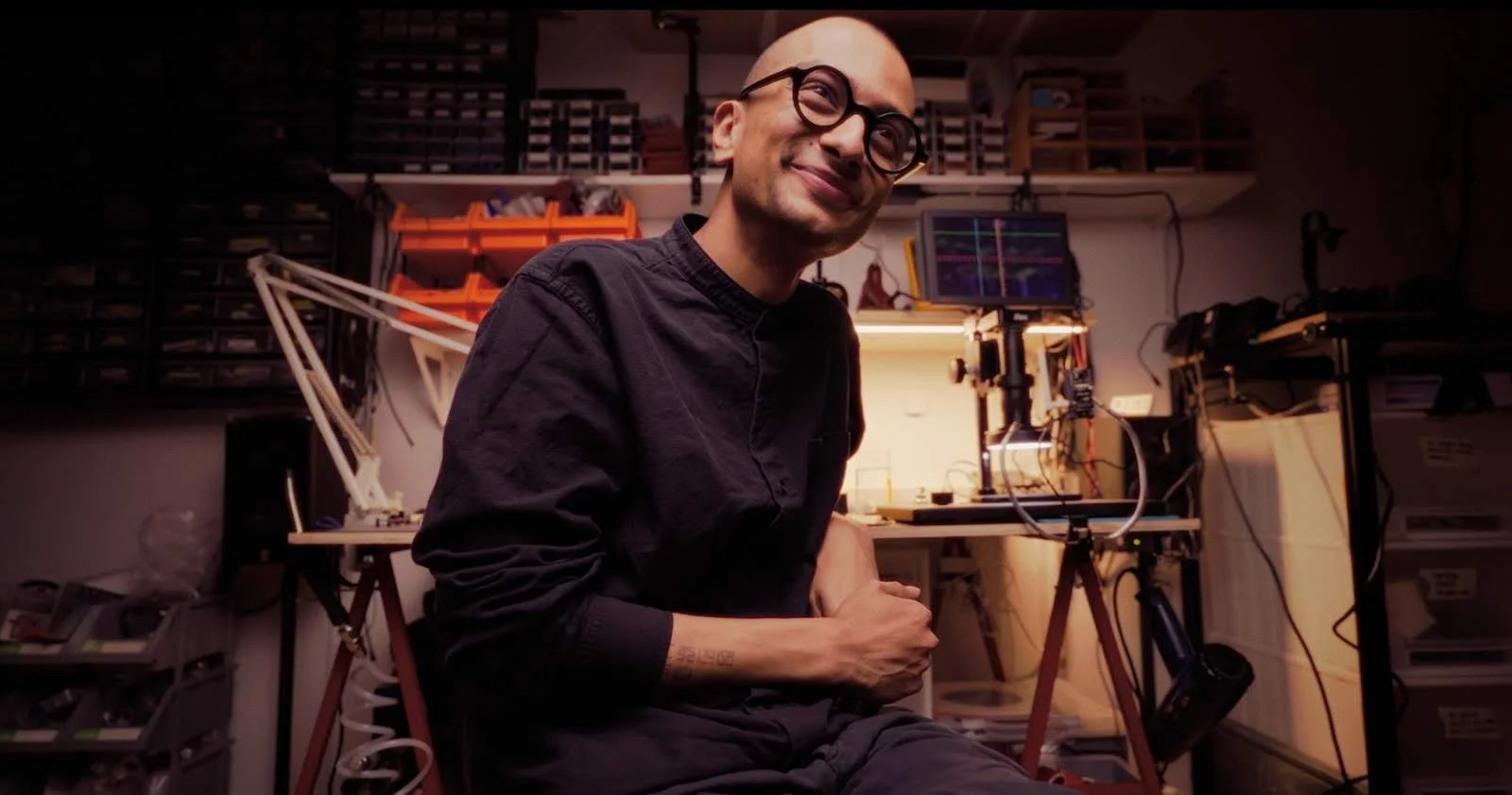 Saurabh Datta sitting in his studio, smiling, surrounded by electronics equipment, component drawers, and workbenches. Warm light from a desk lamp illuminates the space behind him.