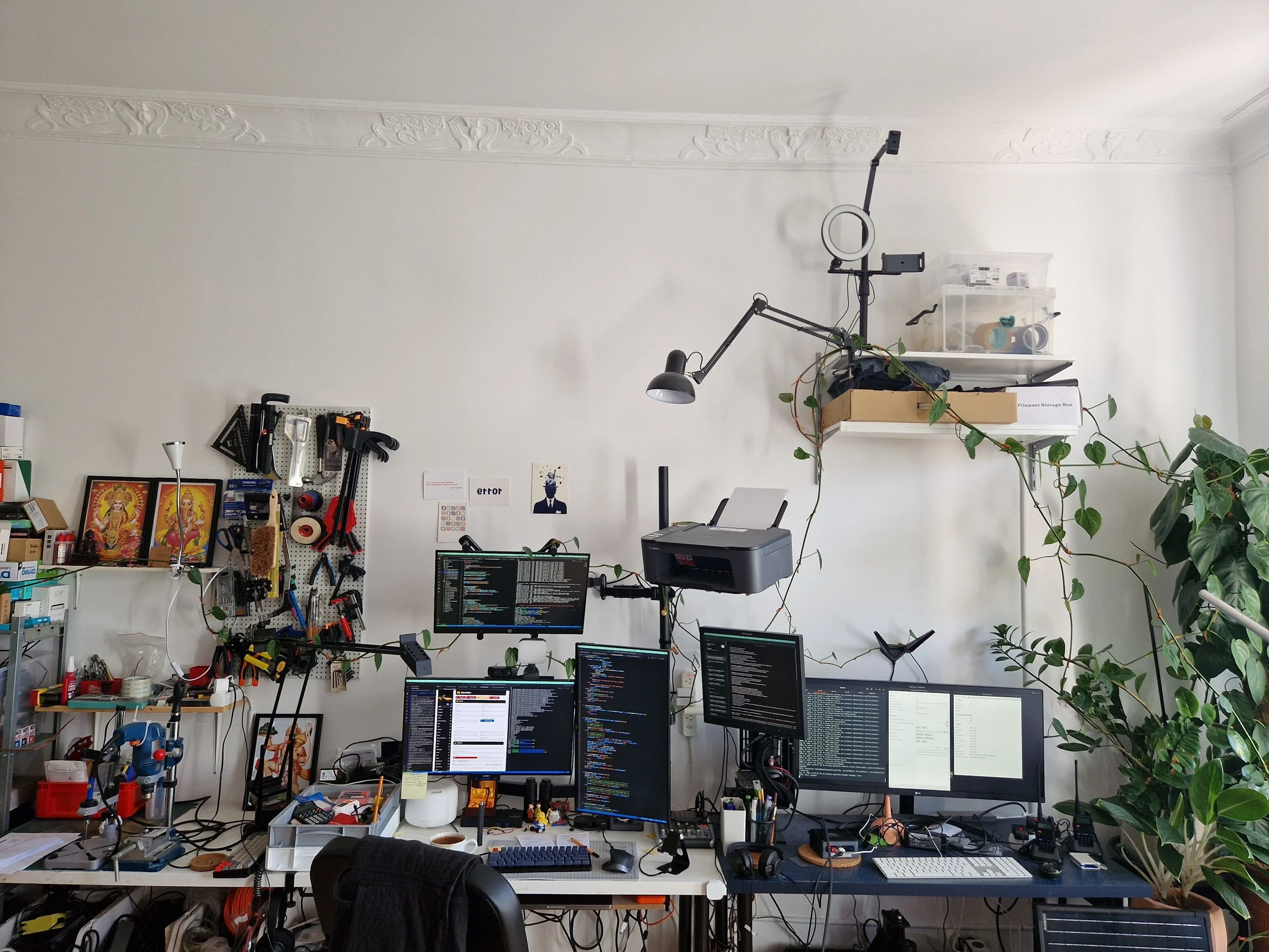 Saurabh Datta's workspace in Berlin — multiple monitors displaying code, a pegboard of tools, trailing plants, and an eclectic mix of objects including small figurines and an "error" print on the wall.
