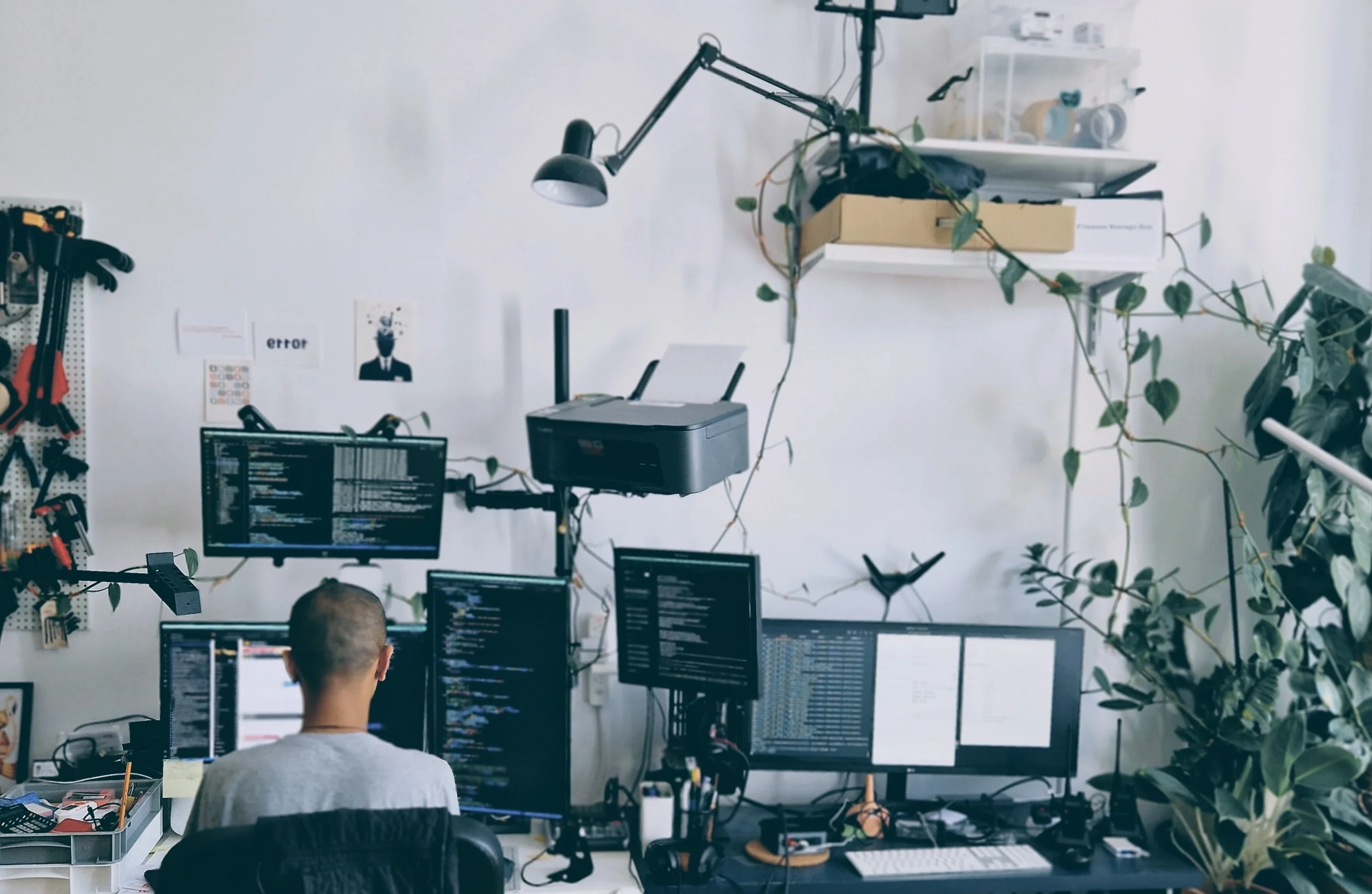 Saurabh Datta seated at a multi-monitor computer workstation in a bright studio, surrounded by coding screens, hanging plants, shelves, tools, and a suspended printer.