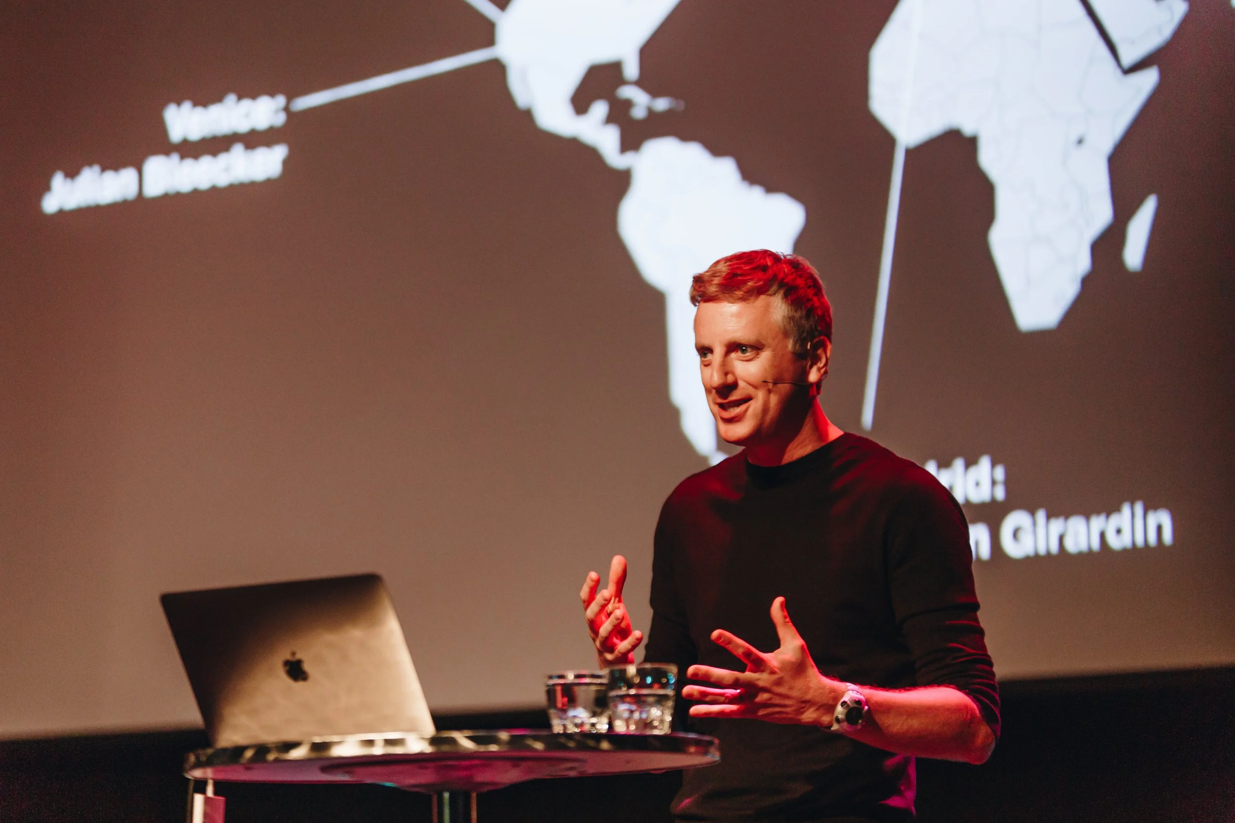 Fabien Girardin presenting on stage, gesturing mid-explanation, with a world map projected behind him showing the Near Future Laboratory's global network. A laptop sits open on the table beside him.