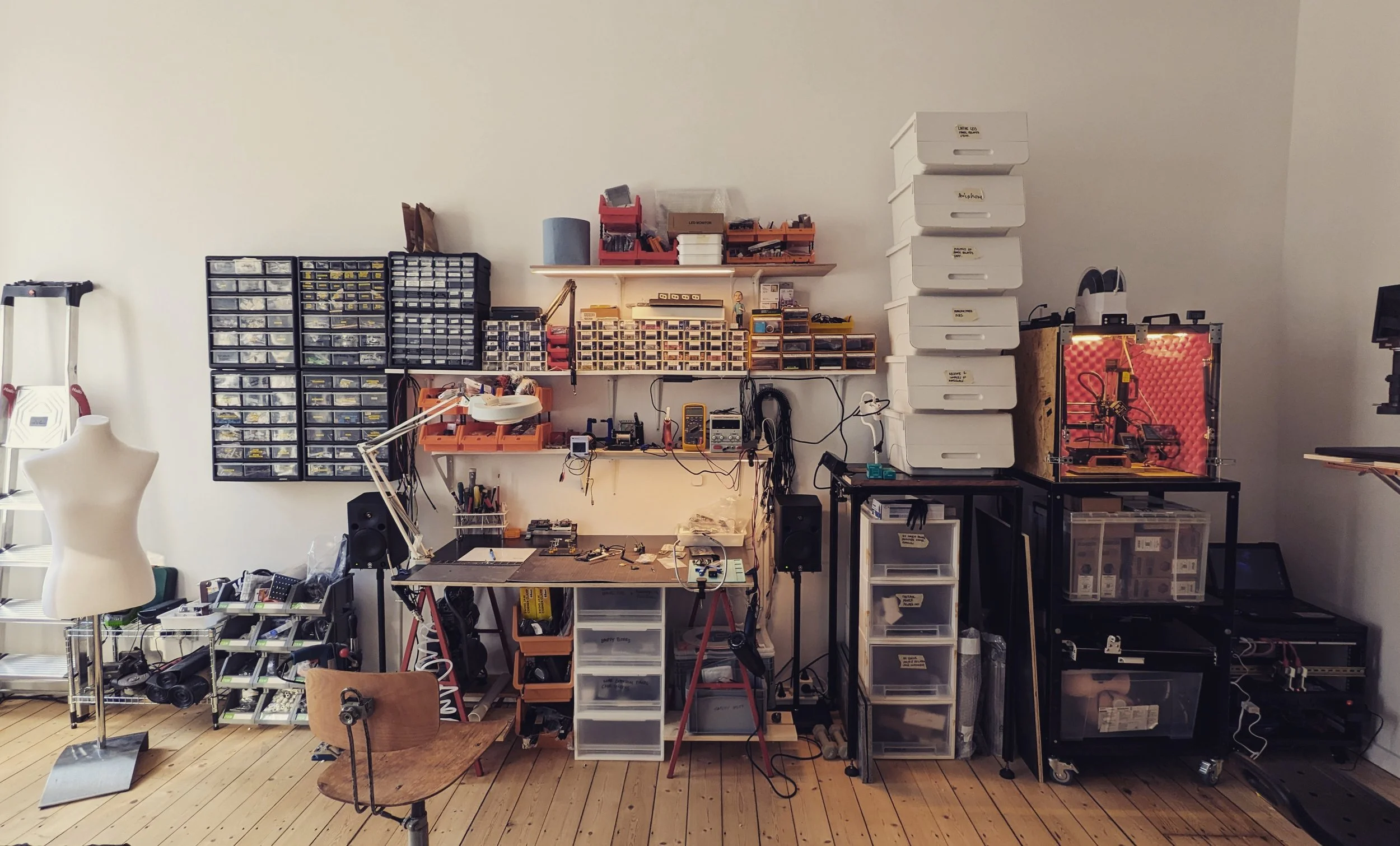 Saurabh Datta's physical prototyping studio — a workbench with soldering equipment, walls lined with labelled component drawers, a dress mannequin, storage boxes, and a 3D printer enclosed in red acoustic foam.