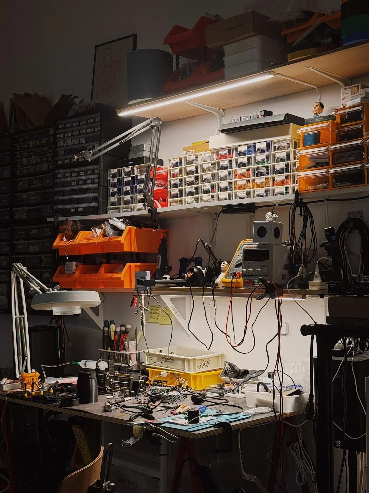Close-up of Saurabh Datta's electronics workbench at night — tangled wires, multimeters, soldering tools, orange parts bins, and rows of small component drawers lit by a strip light overhead.