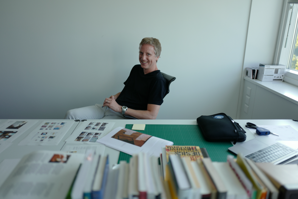Fabien Girardin at his desk, smiling, surrounded by printed layouts, design books, and a cutting mat — a workspace that bridges research, editorial work, and hands-on making.