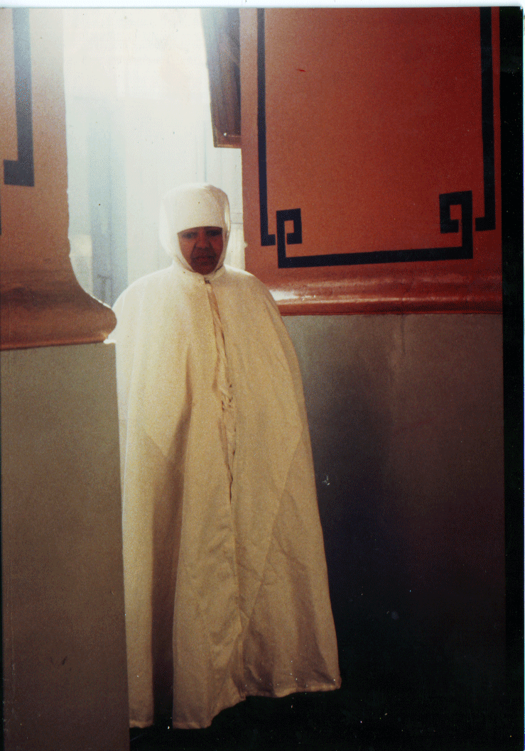 A person dressed in traditional white religious clothing, including a long robe and head covering, standing inside a room with patterned wall decorations.