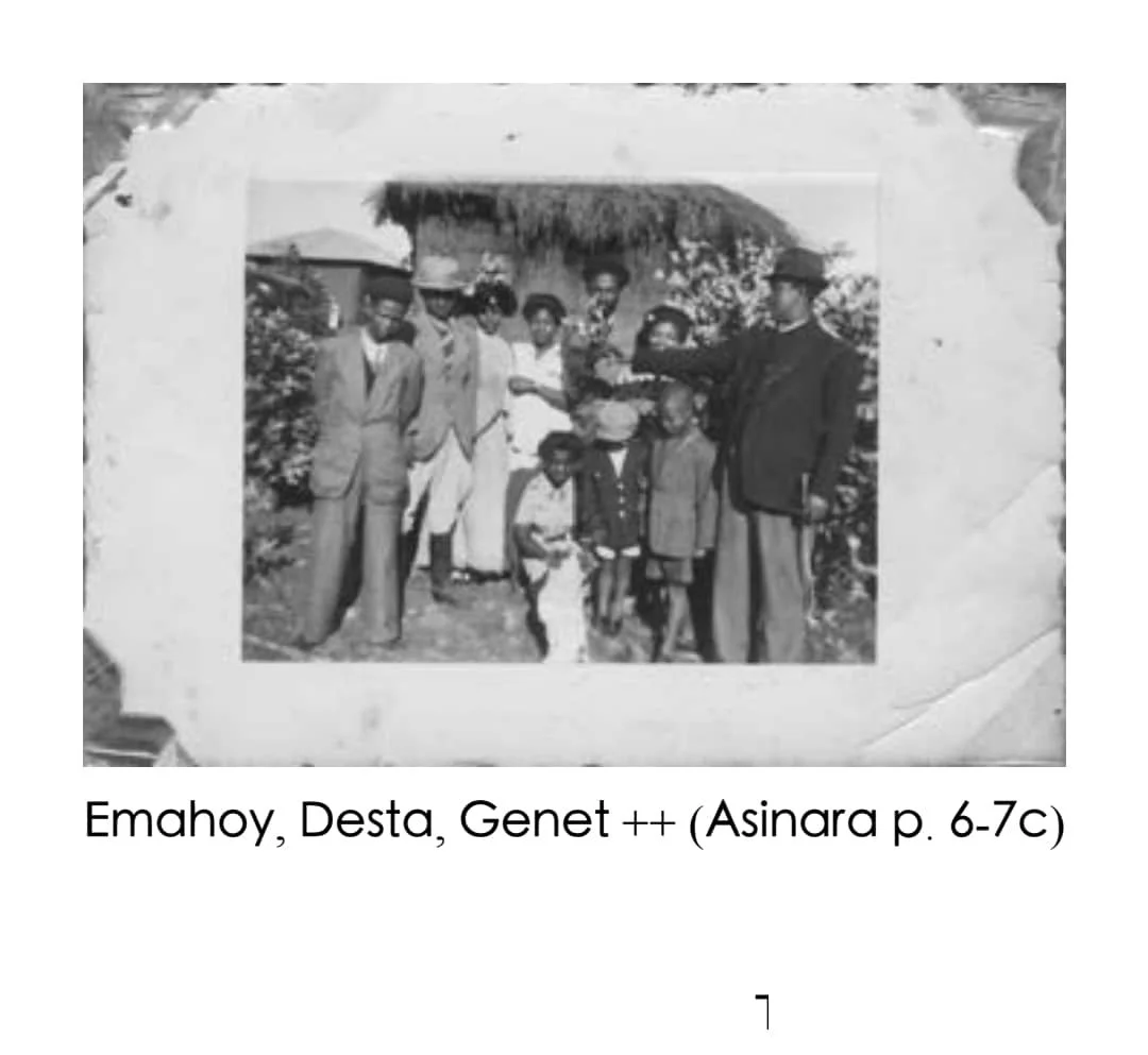 A black and white photo of several people standing outdoors, including men, women, and children, some in traditional and some in Western clothing, against a background of a thatched roof hut and foliage.