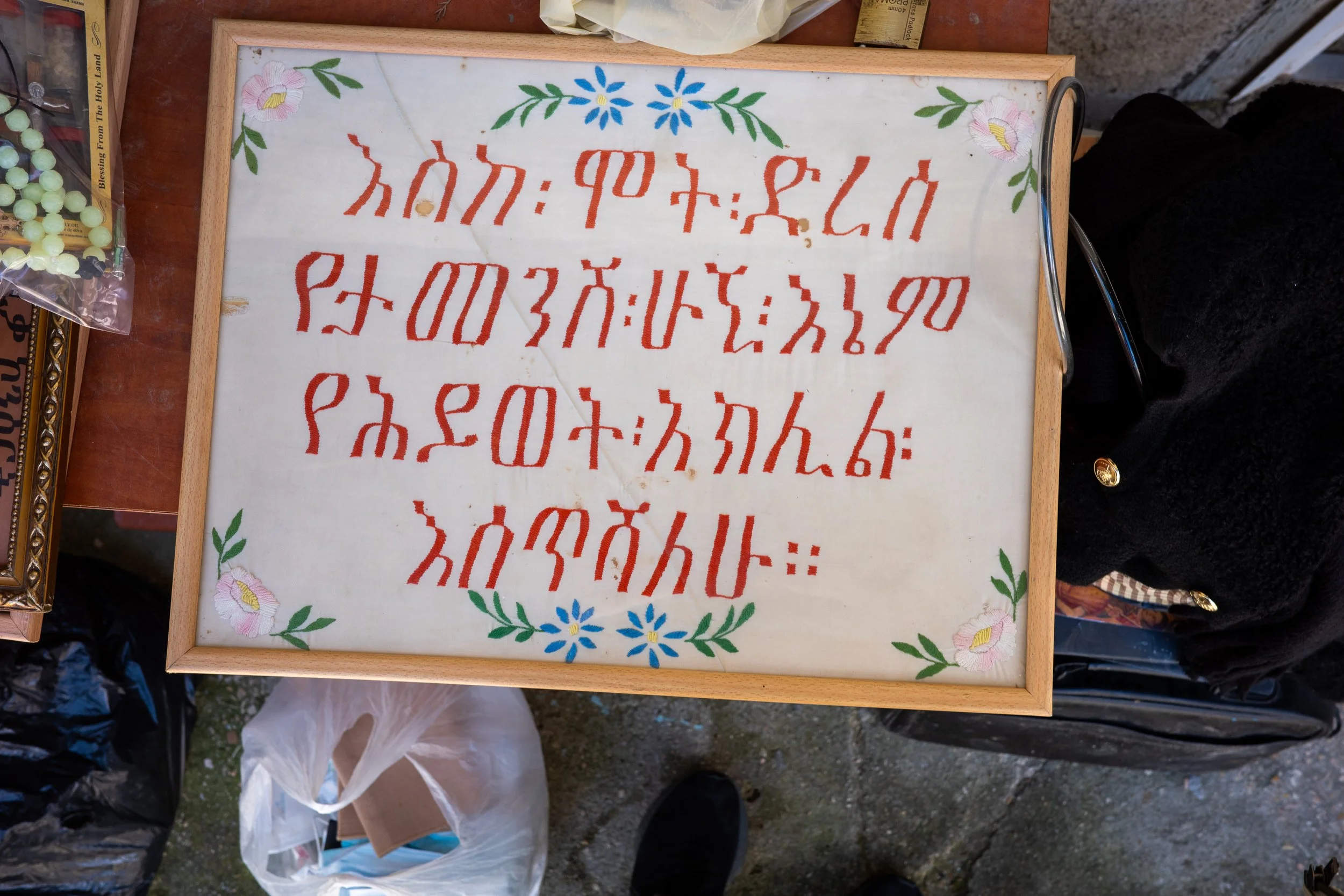 A framed decorative piece with Hawaiian alphabet characters written in red on a white background, adorned with pink and purple flowers and green leaves in the corners.