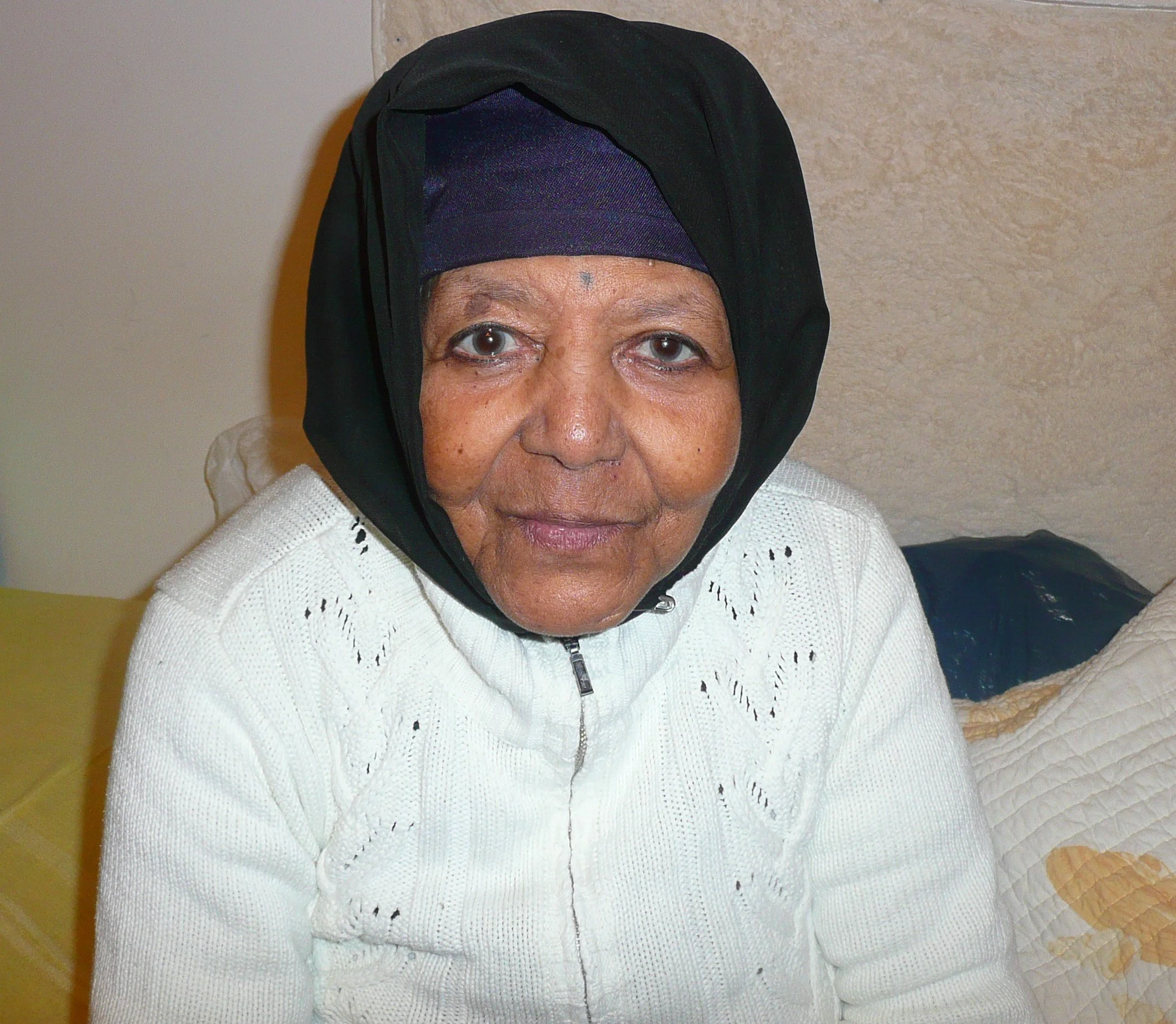 Close-up of an elderly woman wearing a black headscarf and a white knit sweater, sitting against a beige wall.