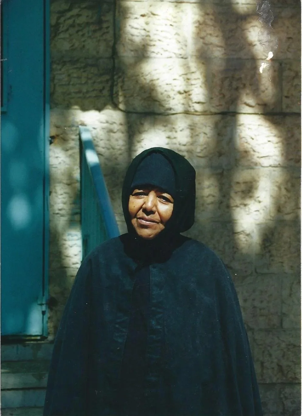A woman wearing a black hooded cloak and face covering, standing outdoors with a stone wall in the background. Shadows and sunlight are cast on her face and the wall.