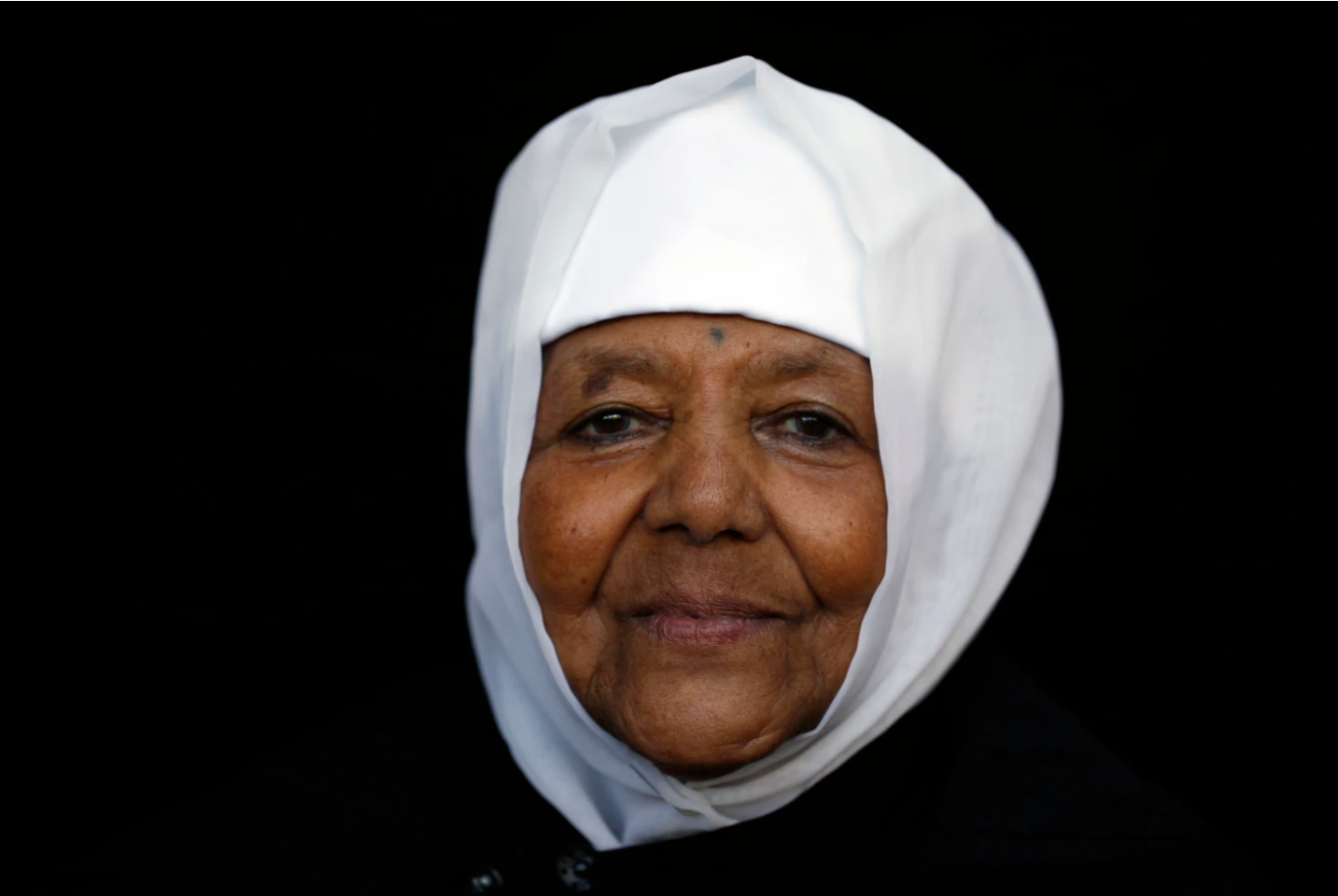 Close-up portrait of an elderly woman wearing a white headscarf against a black background.