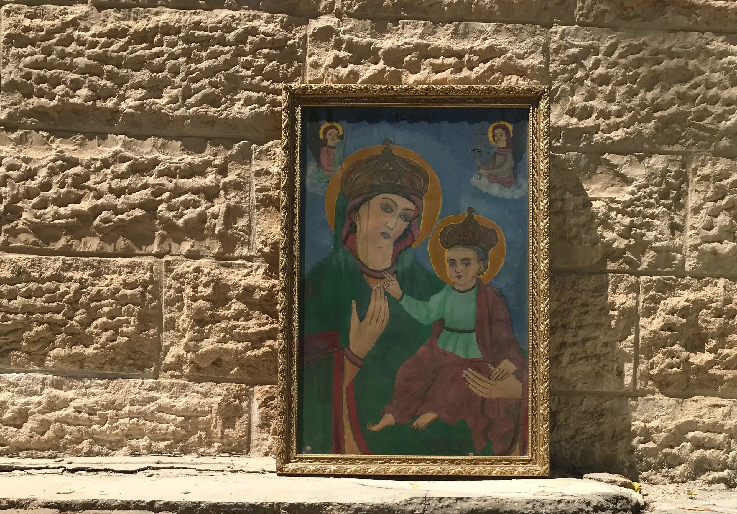A religious painting of a woman with a crown and a halo, holding a child with a halo, against a blue background with clouds and smaller figures. The painting is framed in ornate gold and mounted on a textured stone wall.