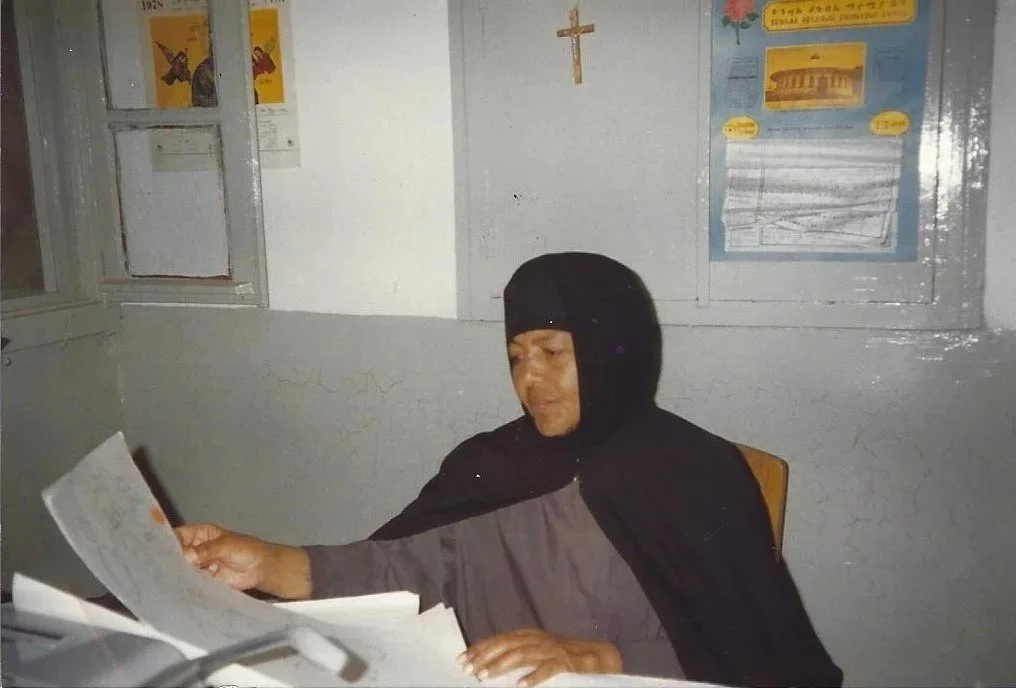 A person wearing a black head covering and gray outfit sitting at a desk, reading papers, with yellow posters and a crucifix on the wall behind them.