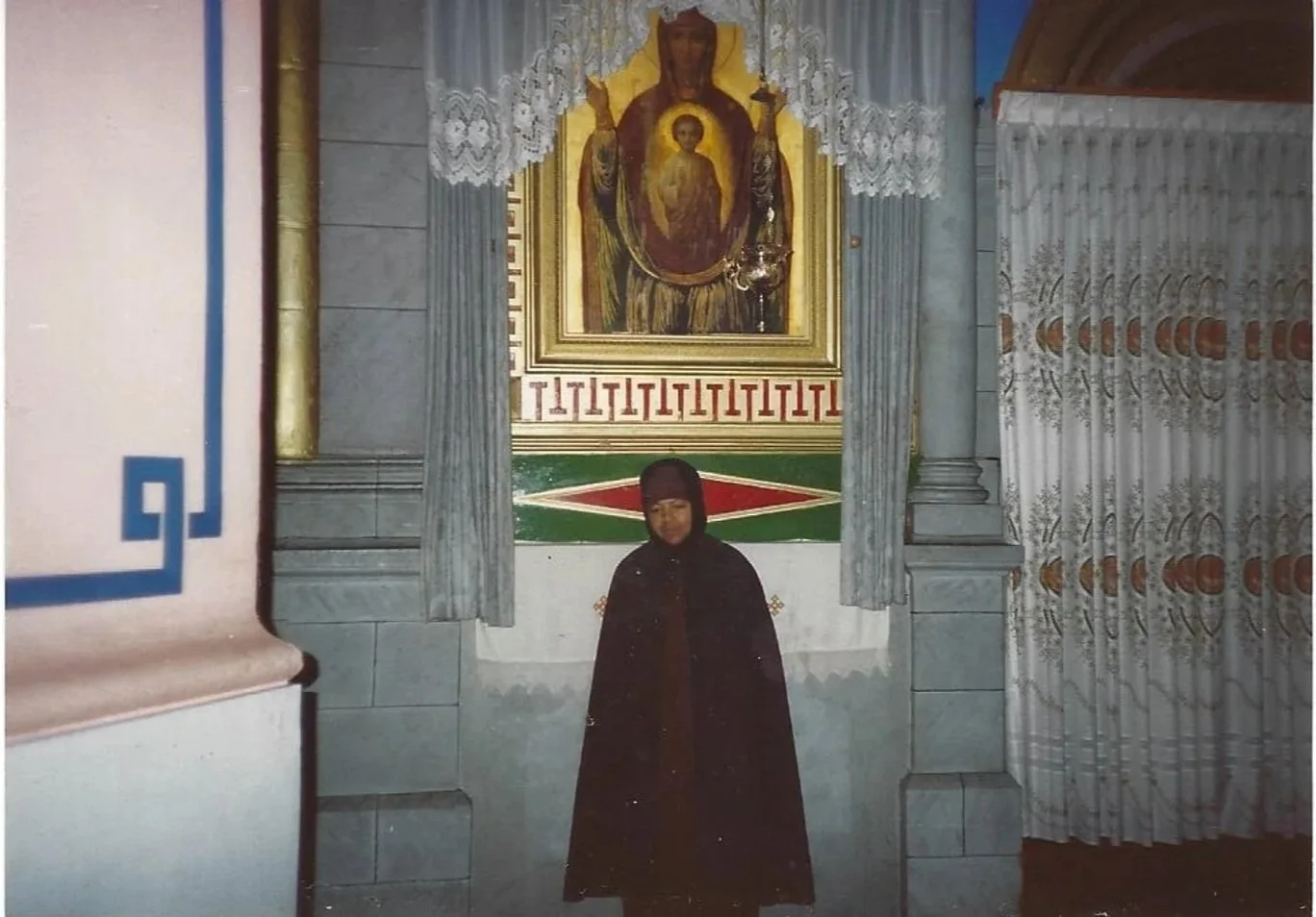 A young girl dressed in a black coat and head covering standing in front of a religious icon of Our Lady of Guadalupe inside a church.