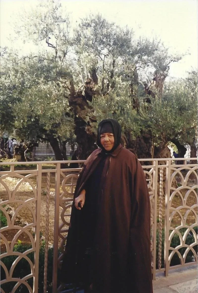 A woman dressed in black with a headscarf standing next to a decorative fence, with large ancient-looking trees in the background.