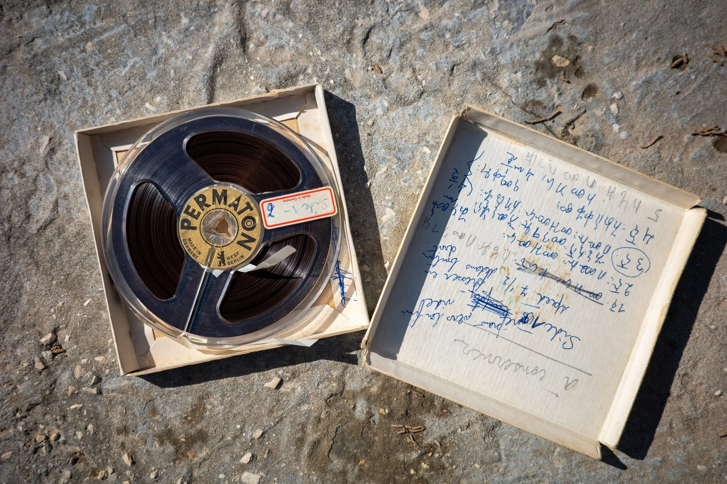 A vintage film reel in a plastic case and a handwritten notebook are placed on a rough concrete surface outdoors.