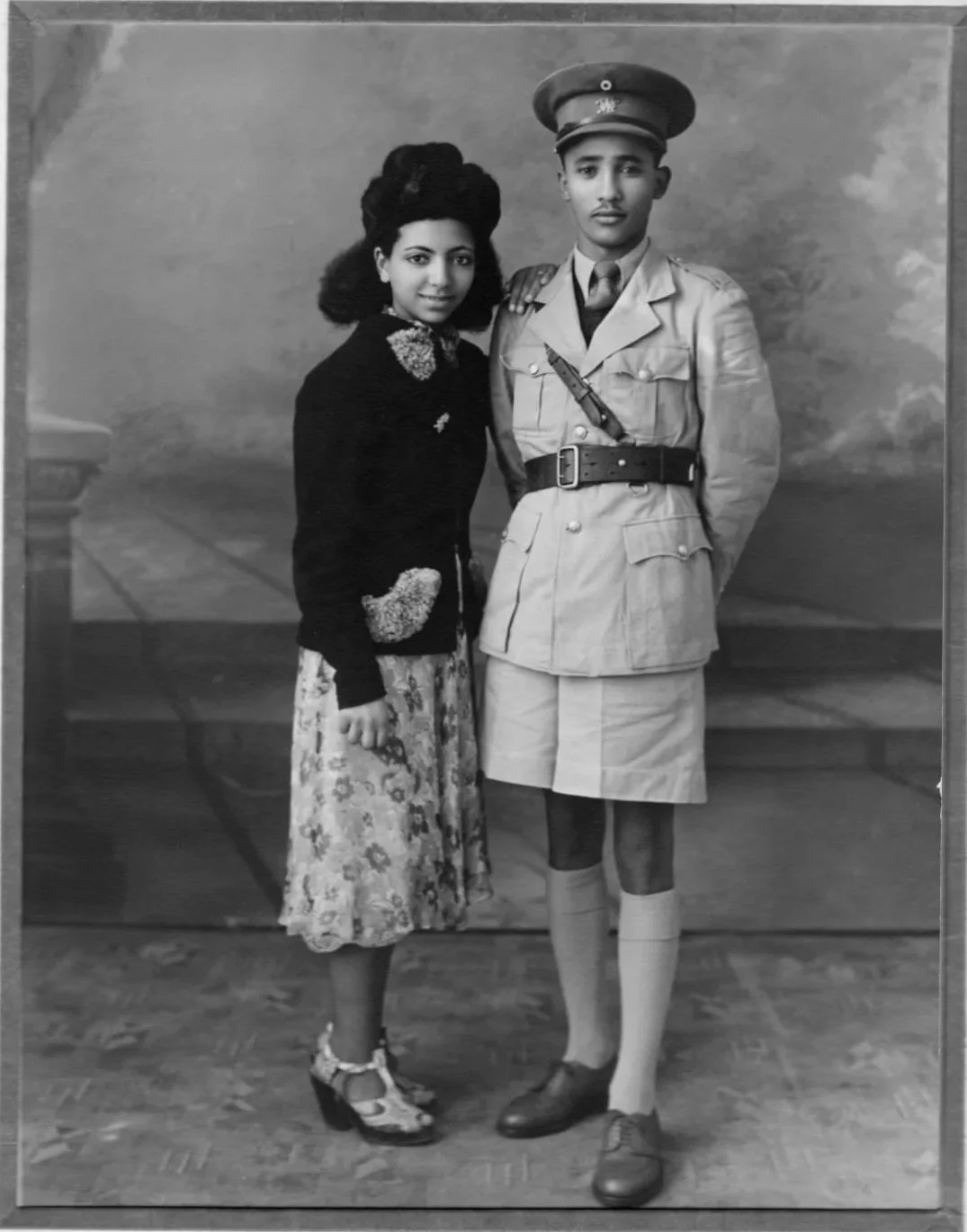 A black-and-white photograph of a young woman and a young man in a studio setting. The woman wears a floral skirt, a dark sweater with decorative details, and heeled shoes. The man wears a military uniform with a cap, belt, and tie.