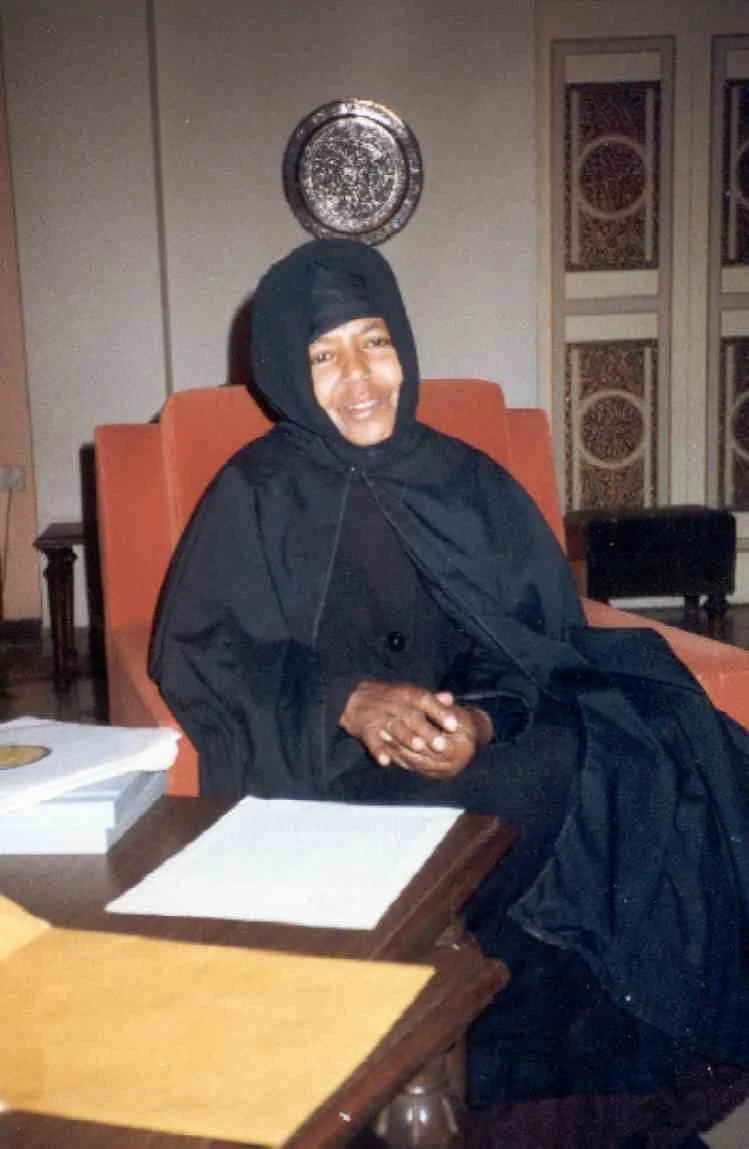 A woman wearing a black outfit and hijab sitting in an orange armchair, smiling with hands clasped, in a room with decor on the walls and a table in front of her.
