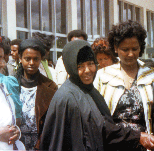 A group of women standing outdoors near a building with large glass windows, one woman in the forefront wearing a black headscarf and coat, smiling at the camera.