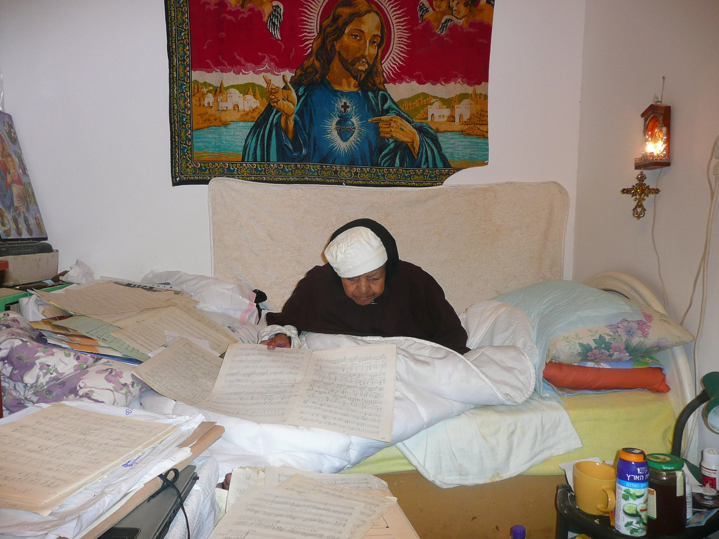 A person with a head covering sitting on a bed surrounded by scattered papers, musical sheets, and books, with religious artwork featuring Jesus on the wall behind.