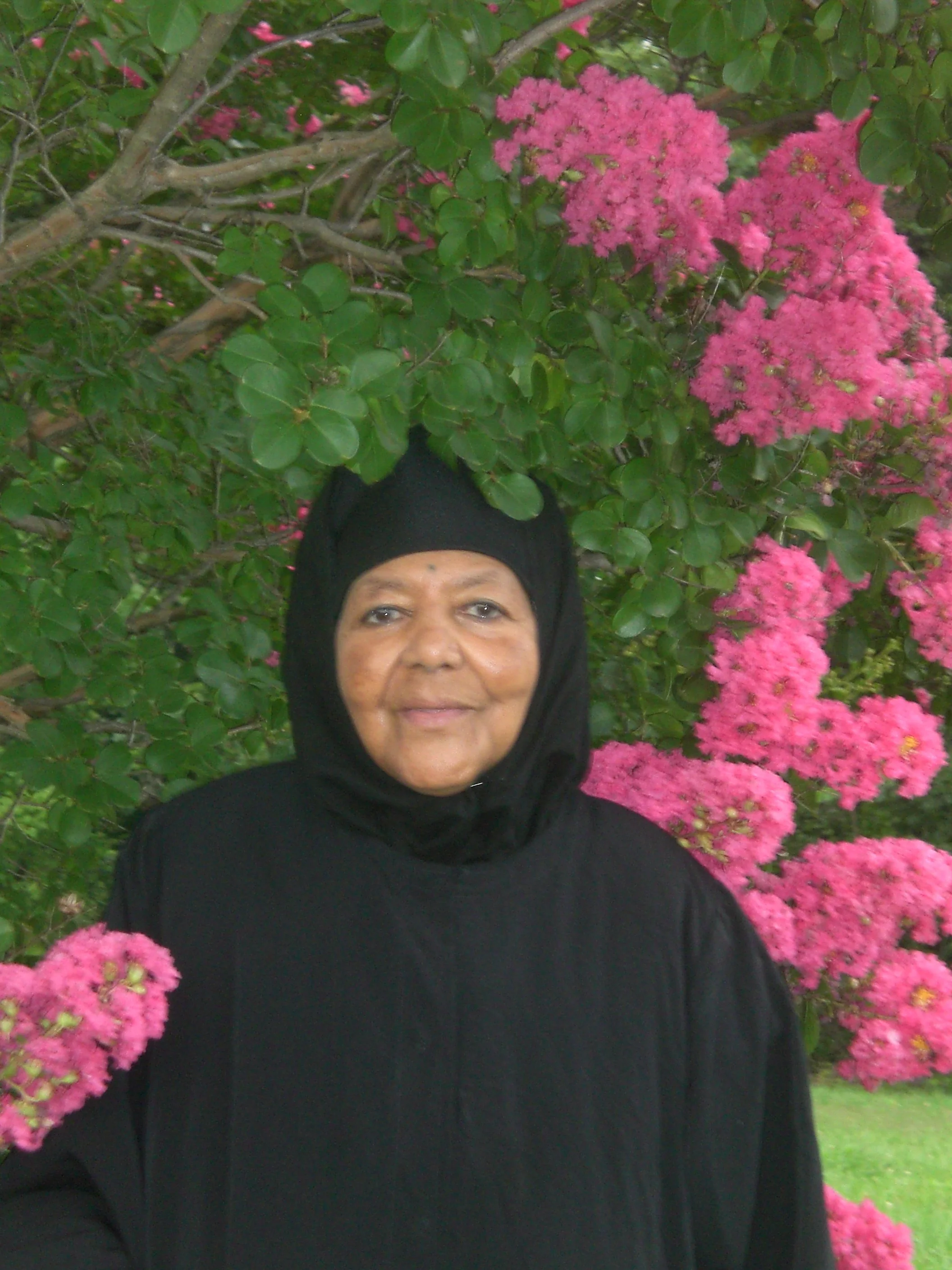 A woman wearing a black hijab and black clothing standing in front of a bush with pink flowers.