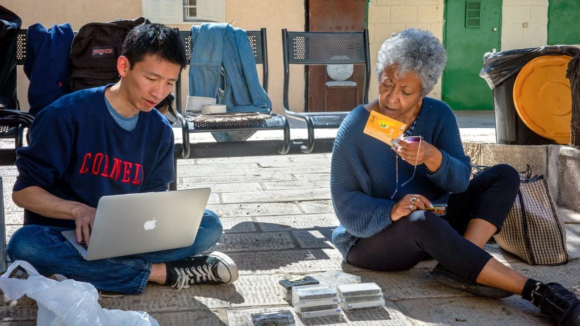 A young man and an elderly woman sitting on the sidewalk, using electronic devices. The young man is working on a laptop, and the elderly woman is listening to something with earphones, with a stack of CDs or DVDs in front of them.