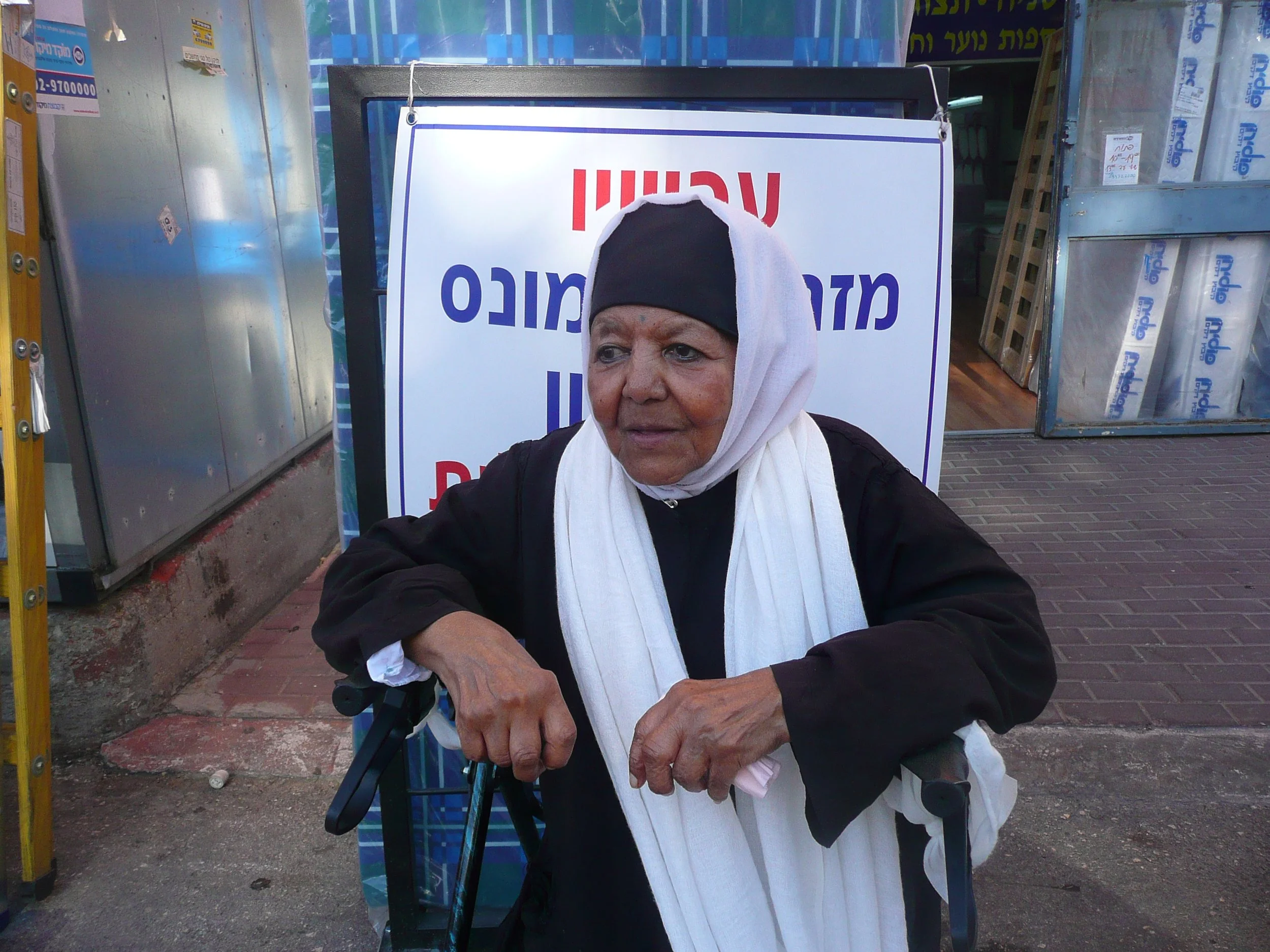 An elderly woman with a headscarf sitting on a wheelchair outside a store, next to a sign with Hebrew writing.