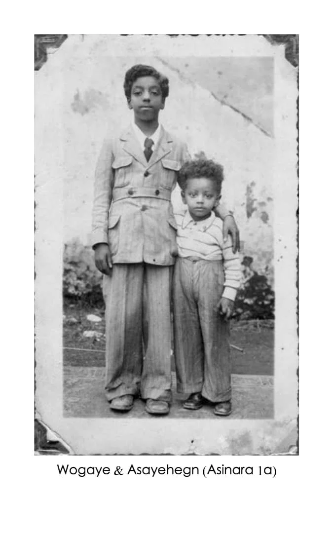 Black and white photo of two young boys standing outdoors, one taller and older with his arm around the shorter younger boy. Both are dressed in vintage clothing. The background shows a wall and some plants.