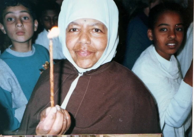 A smiling woman wearing a white headscarf and brown clothing holding a lit birthday candle, surrounded by children.