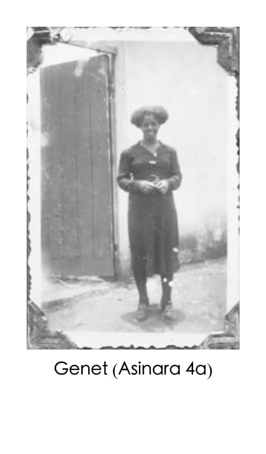 A black-and-white photo of a woman standing outdoors near a building with a wooden door open behind her. She is wearing a dress and has short, curly hair, and is smiling while holding her hands together in front of her.