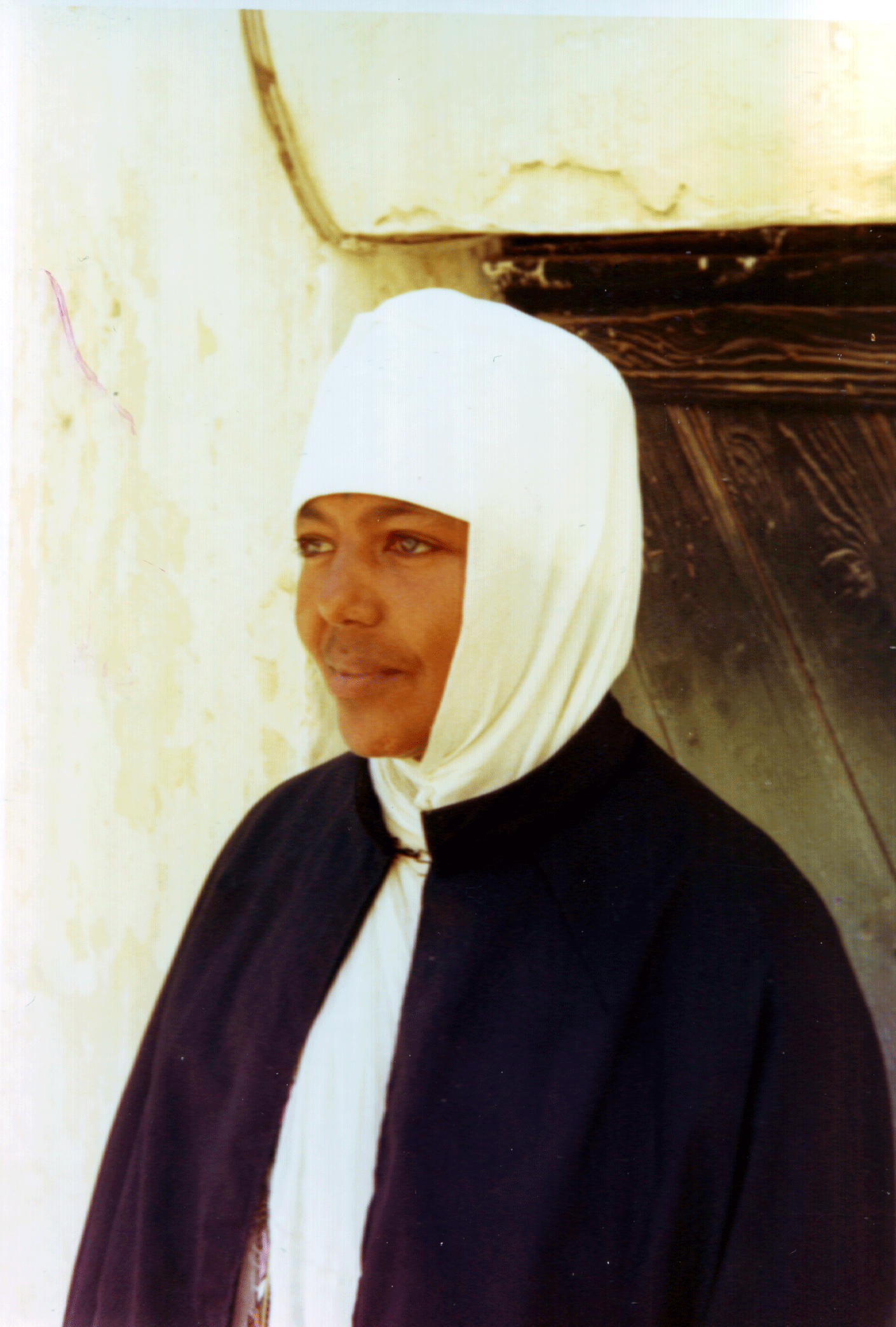 Portrait of woman with light eyes and fair skin wearing a white headscarf and black clothing, standing against a yellowish wall with black wooden beams.
