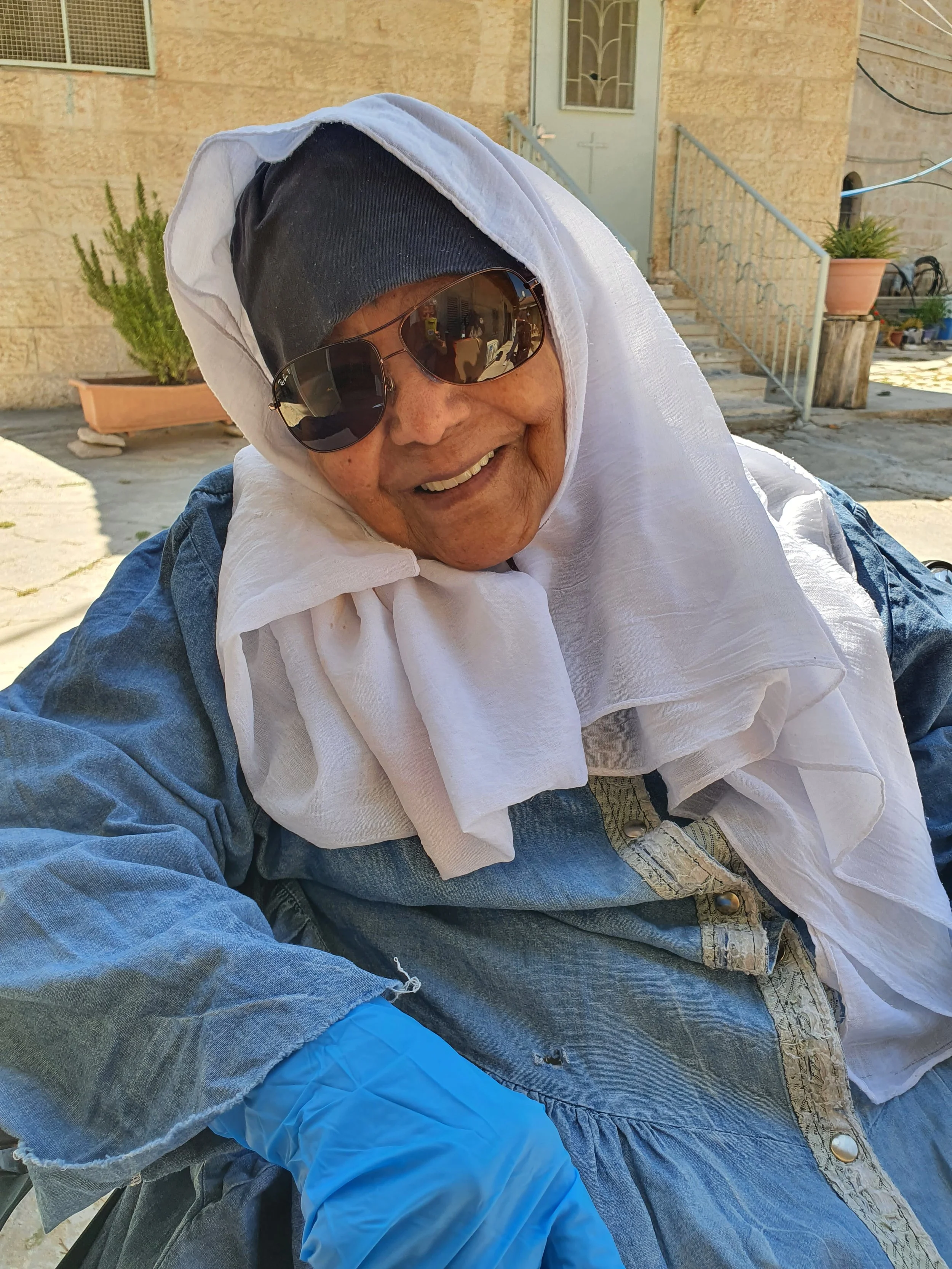 A smiling older woman wearing dark sunglasses, a black headscarf, a white scarf, and a denim jacket outside near a stone building with stairs, potted plants, and a metal railing.