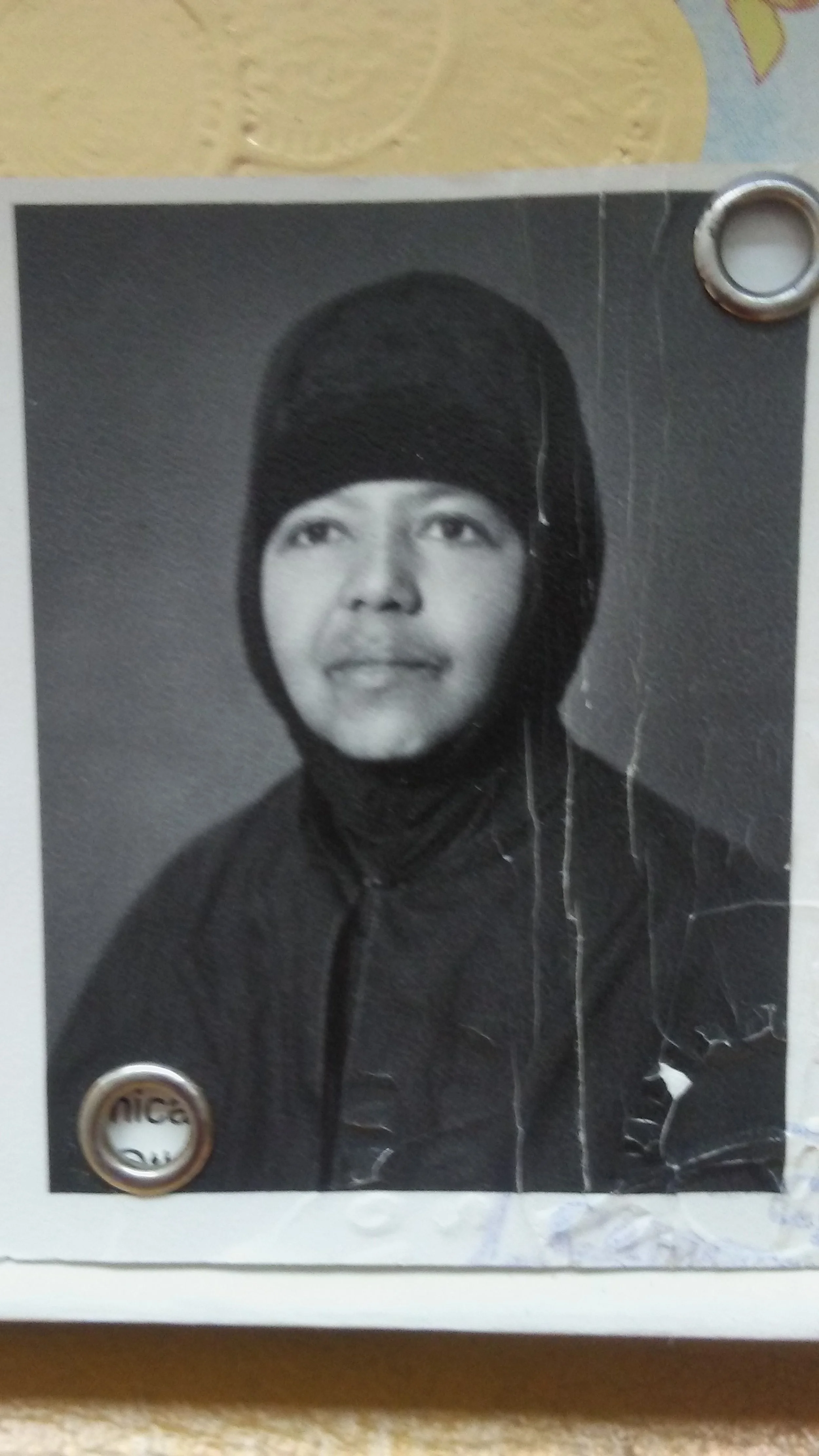 A black and white portrait photograph of a woman wearing a headscarf, mounted on a white background with a metal pin in the top right corner.
