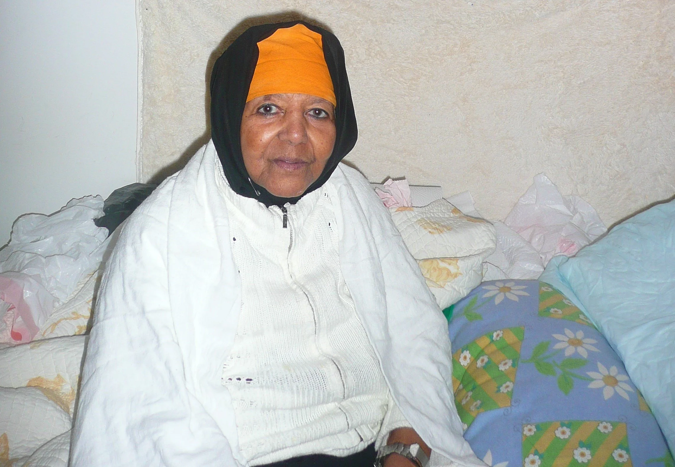 An elderly woman is sitting on a bed, wearing an orange headscarf, a black head covering, a white jacket, and a white knit sweater. The bed has patchwork floral and geometric sheets and pillows. The background features a plain, light-colored wall.