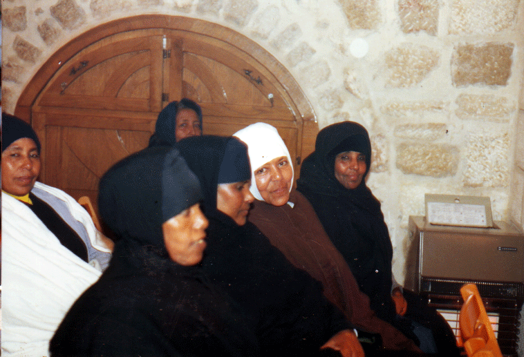 A group of women sitting in a room with stone walls and a wooden door in the background, some wearing headscarves.