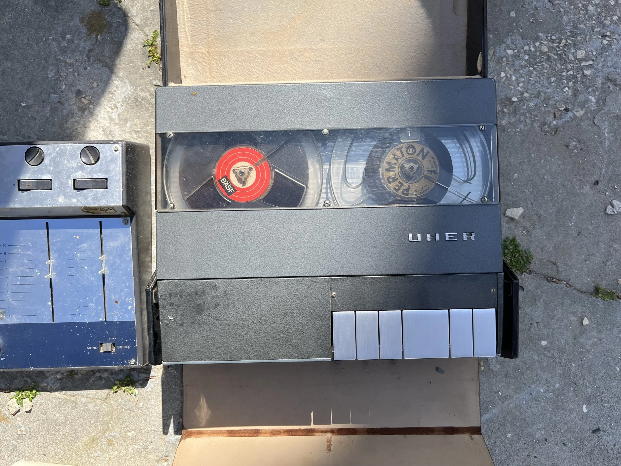 Vintage UHER tape recorder with two reels, one labeled 'Basf' and the other labeled 'Permatone,' positioned on a concrete floor near a small, rusty metal structure and a blue panel with sliders.