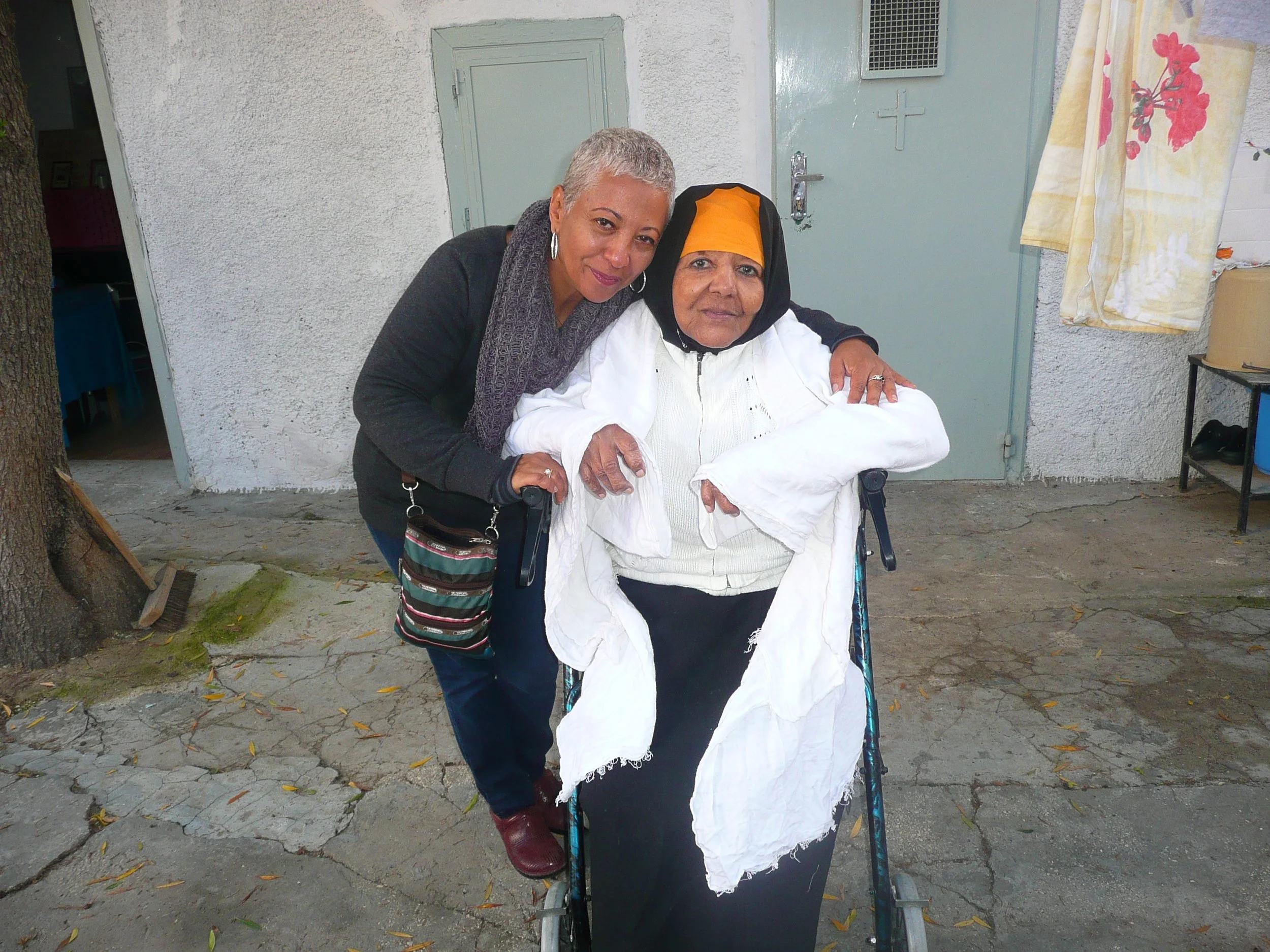 Two women, one elderly and one middle-aged, standing outdoors in front of a light green door and wall, smiling for the photo. The elderly woman is seated in a wheelchair wearing a headscarf, and the middle-aged woman has short gray hair and is leanin